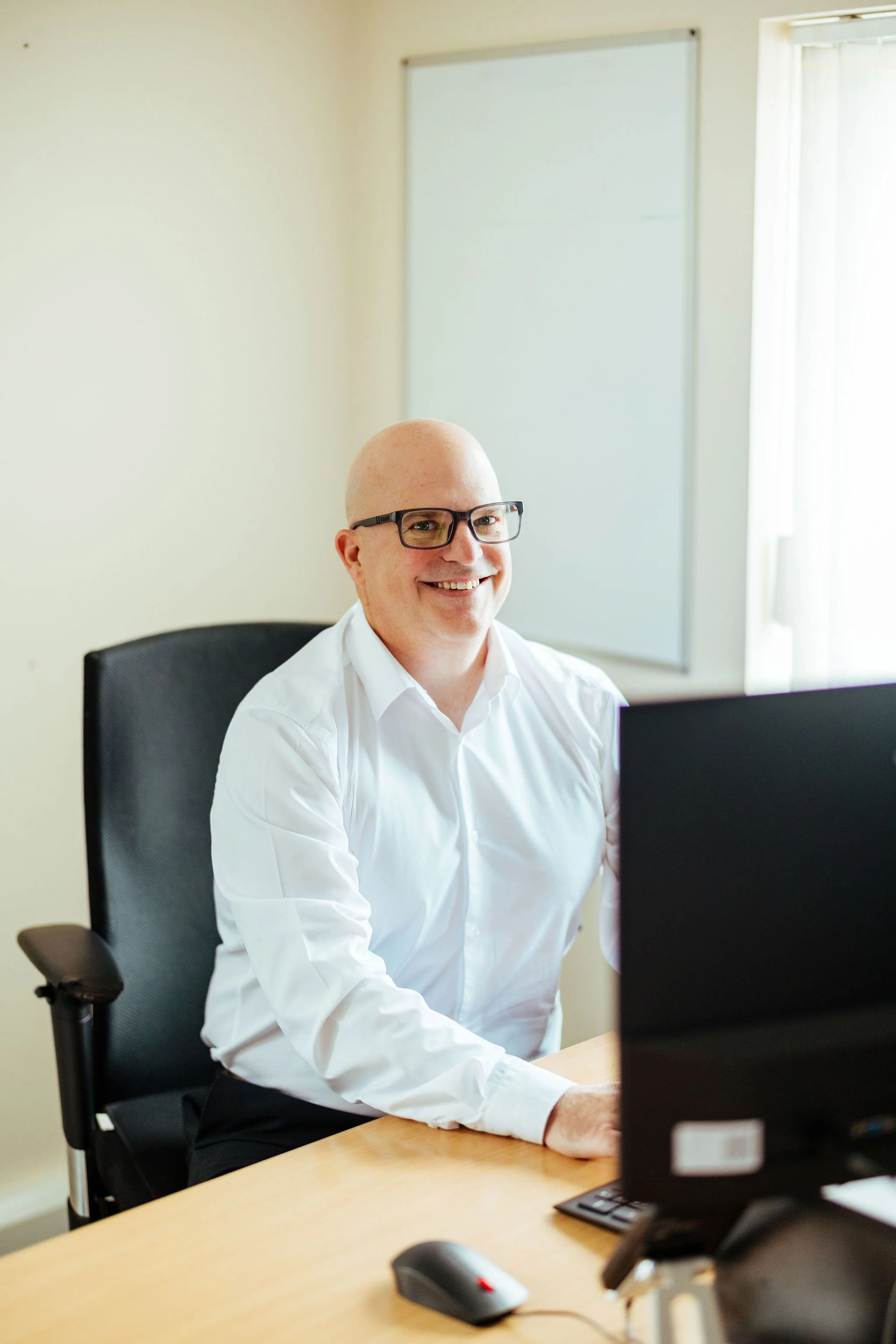 Bald man wearing glasses and a white dress shirt sitting at a desk in front of two computer monitors, smiling at the camera.