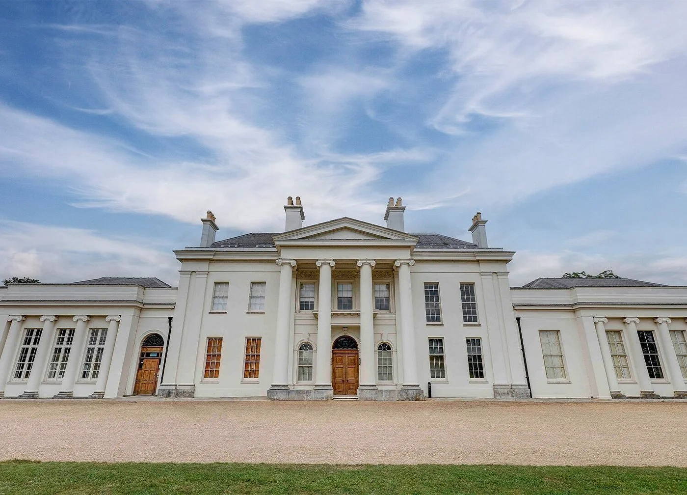 Large white mansion with classical architecture featuring columns, multiple windows, and multiple chimneys under a blue sky with wispy clouds.