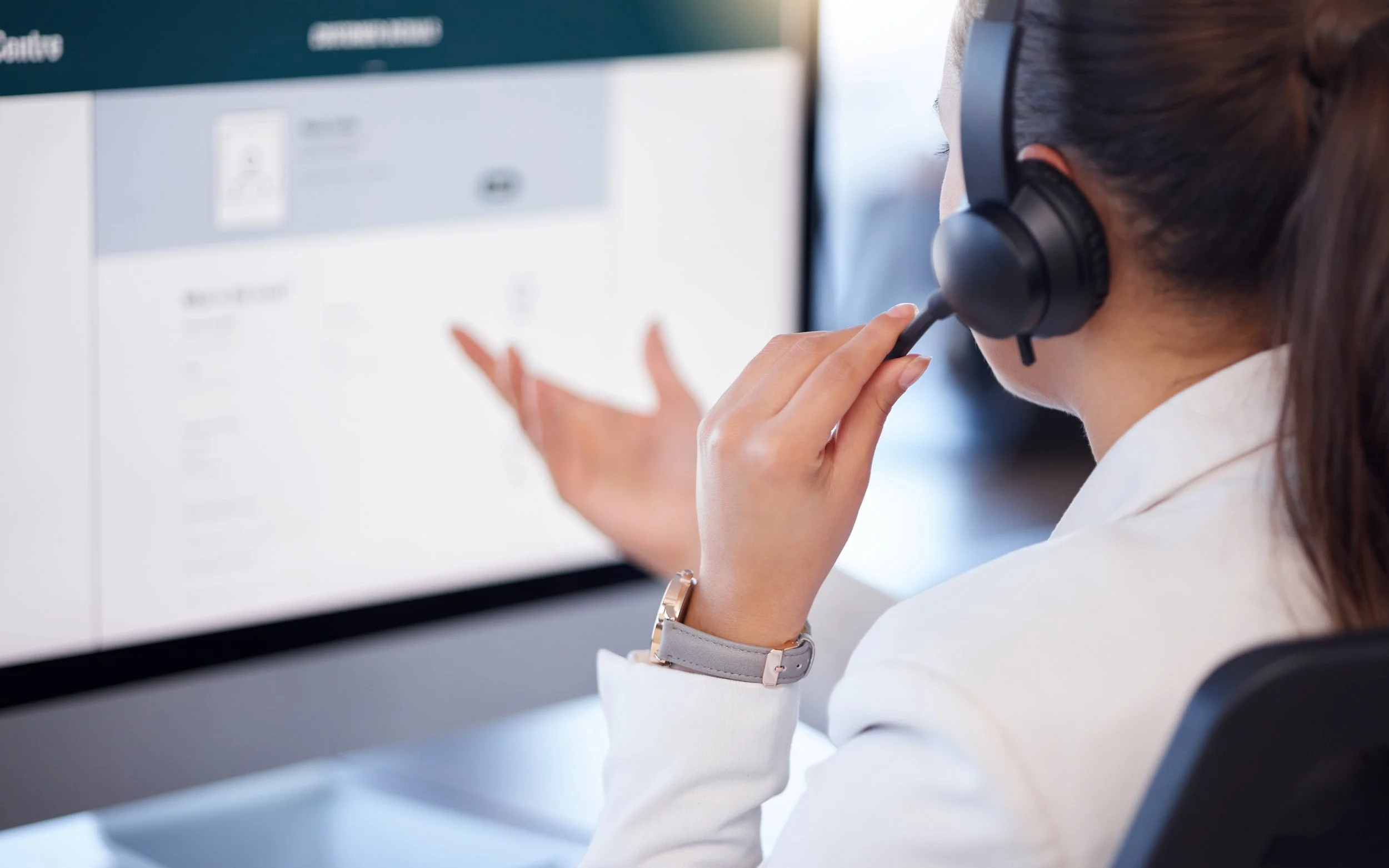 A woman with brown hair in a ponytail wearing a headset and white blazer, sitting at a desk, pointing at a computer screen, with a blurred digital interface visible.