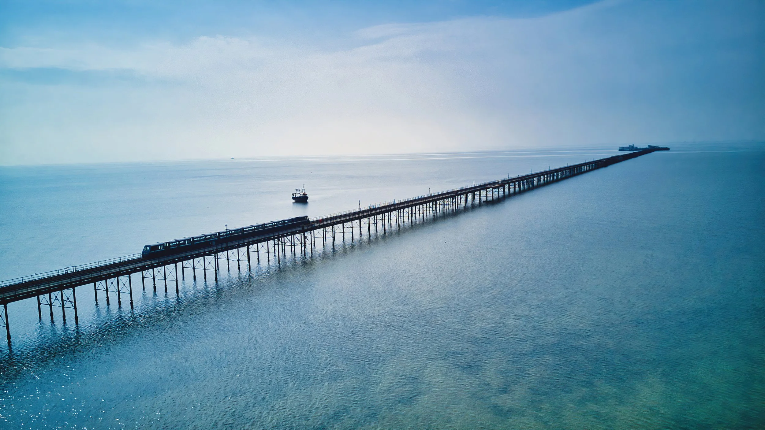 A long pier extends into the ocean with a train on it, a boat nearby, and a building at the end of the pier, under a partly cloudy sky.