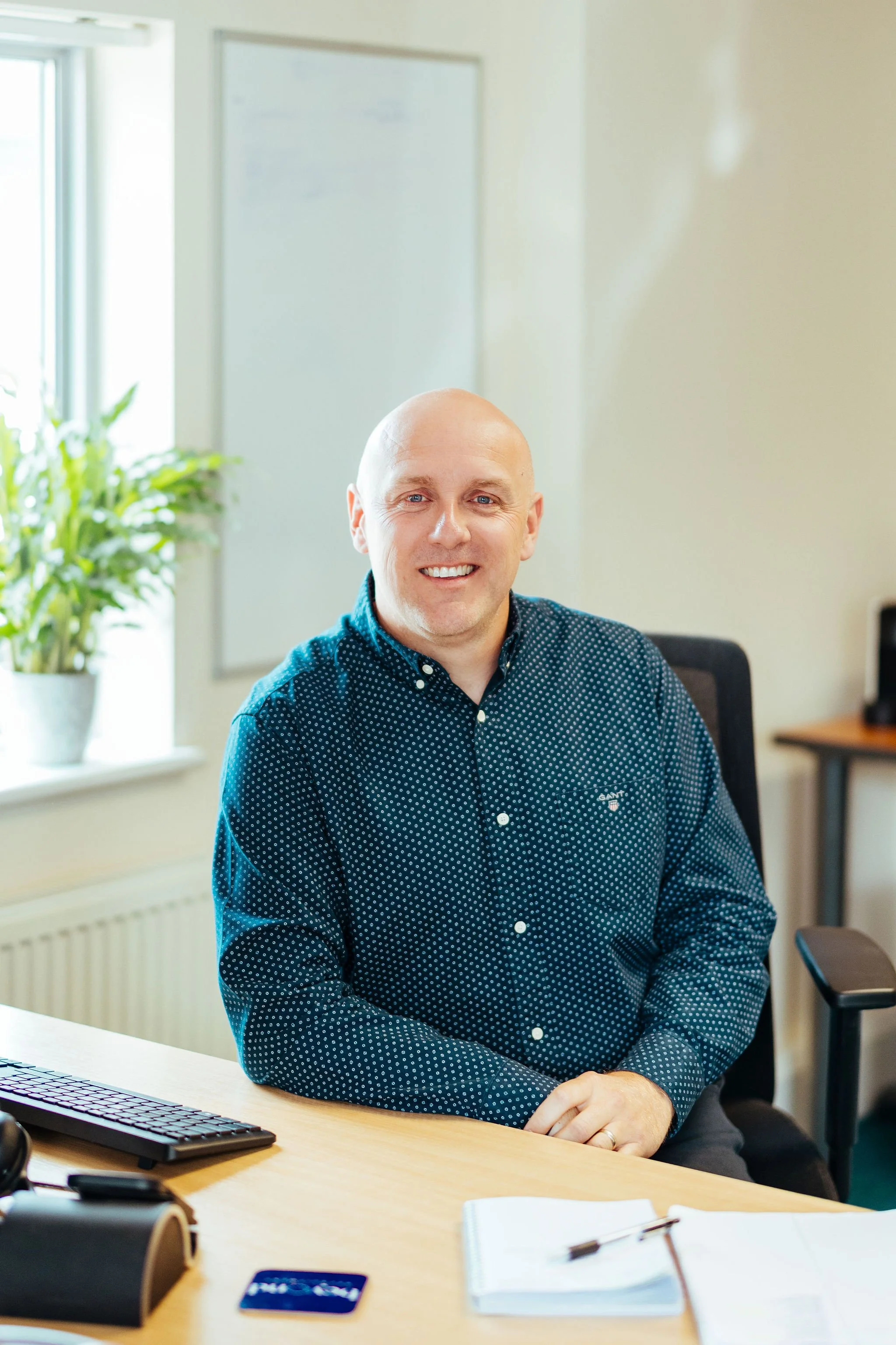 Smiling man sitting at a desk in an office, dressed in a navy polka dot shirt. Office environment with a computer keyboard, smartphone, and office supplies on the desk, with a window and whiteboard in the background.