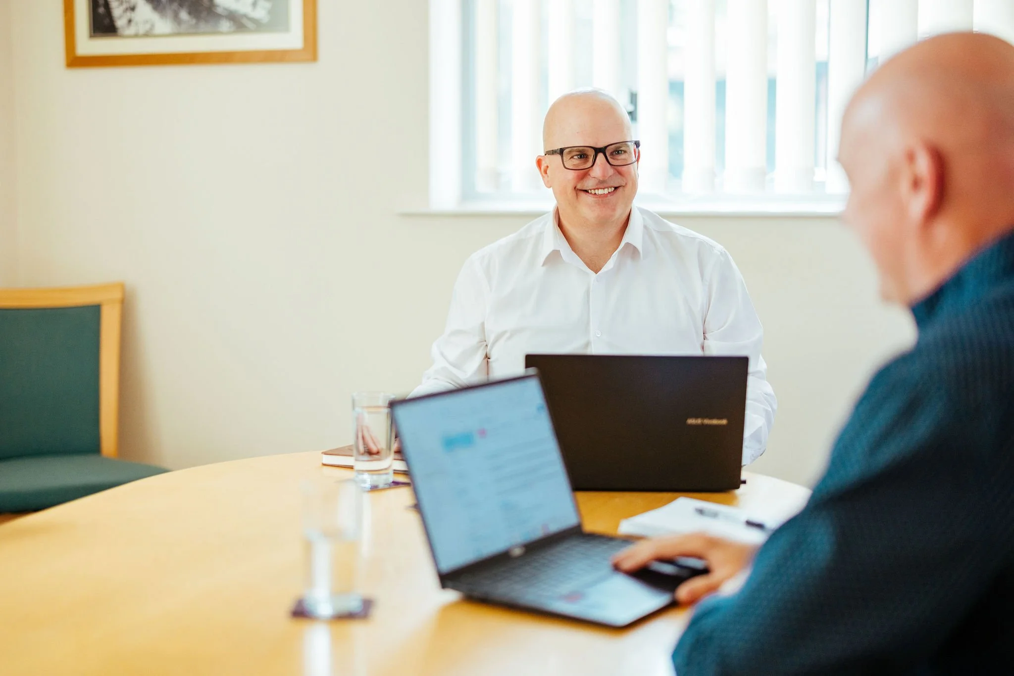 Two men with laptops conversing in a bright office room with a window and a framed picture on the wall.