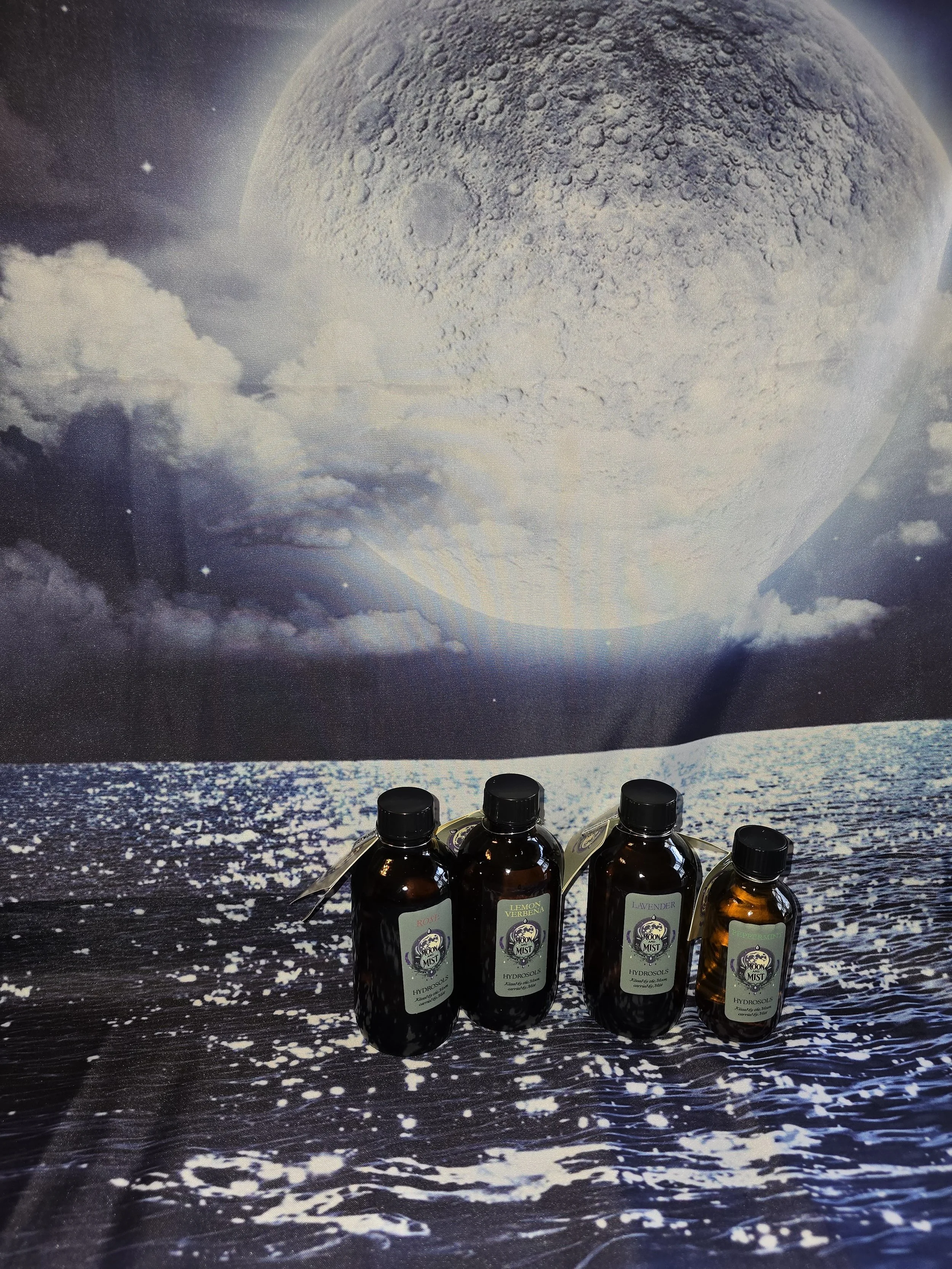 Four amber bottles of hydrosols labeled Lemon Verbena, Rose, Lavender, and another with an indistinguishable label, placed on a surface with a backdrop that appears to be a black and white photo of the Moon and the ocean.