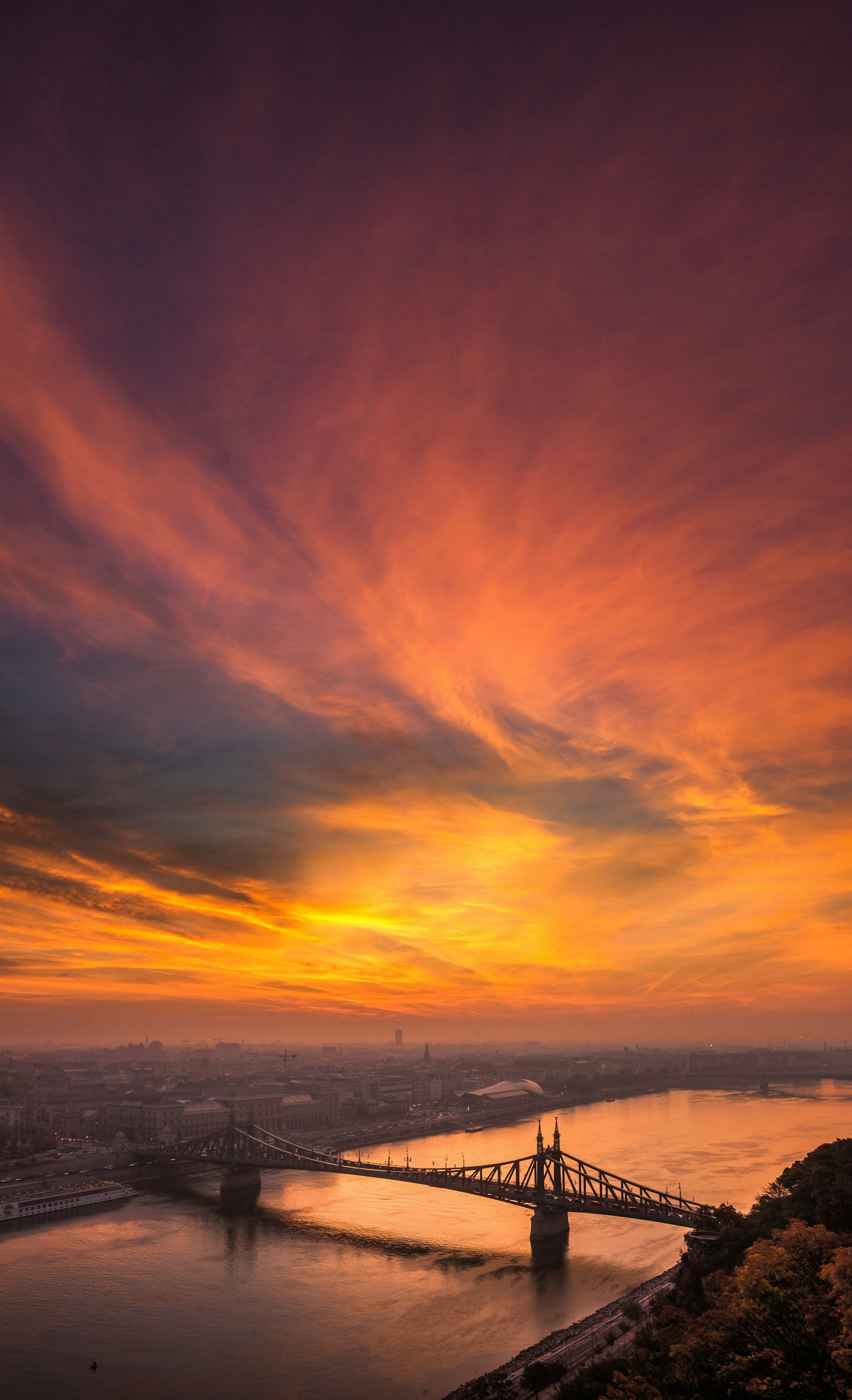 A cityscape during sunset with colorful clouds in the sky, a bridge crossing a river, and buildings along the riverbank.