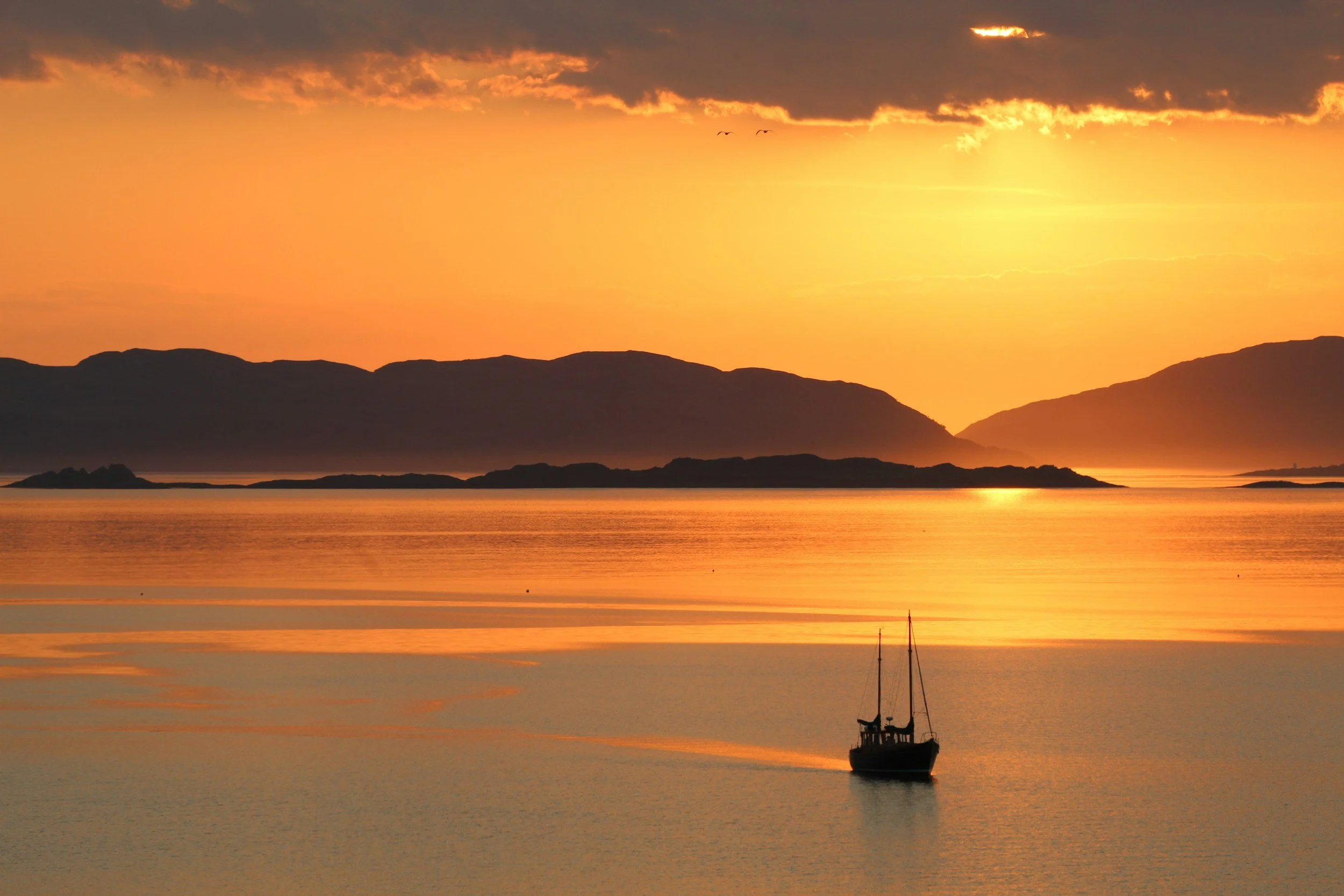 Sunset over calm water with a silhouette of a sailboat and distant mountains.