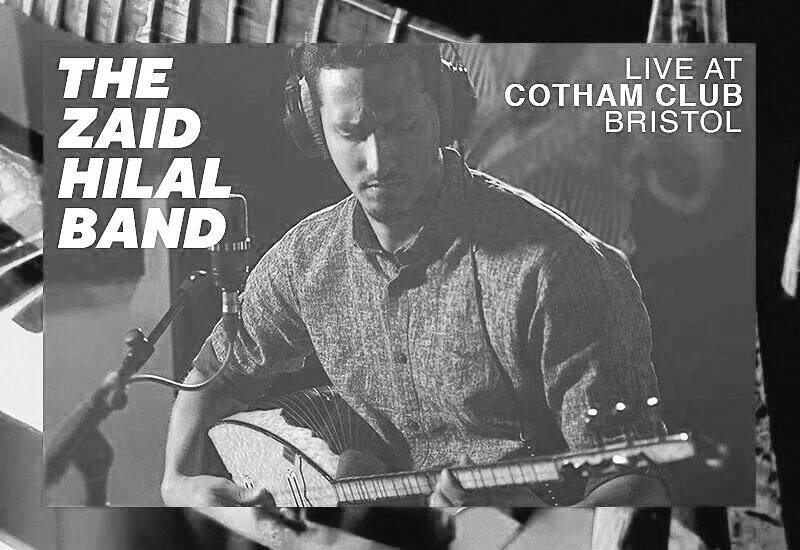 A man in a gray shirt playing an acoustic guitar with a microphone in front of him, performing live at Cotham Club Bristol, promoting The Zaid Hilal Band.