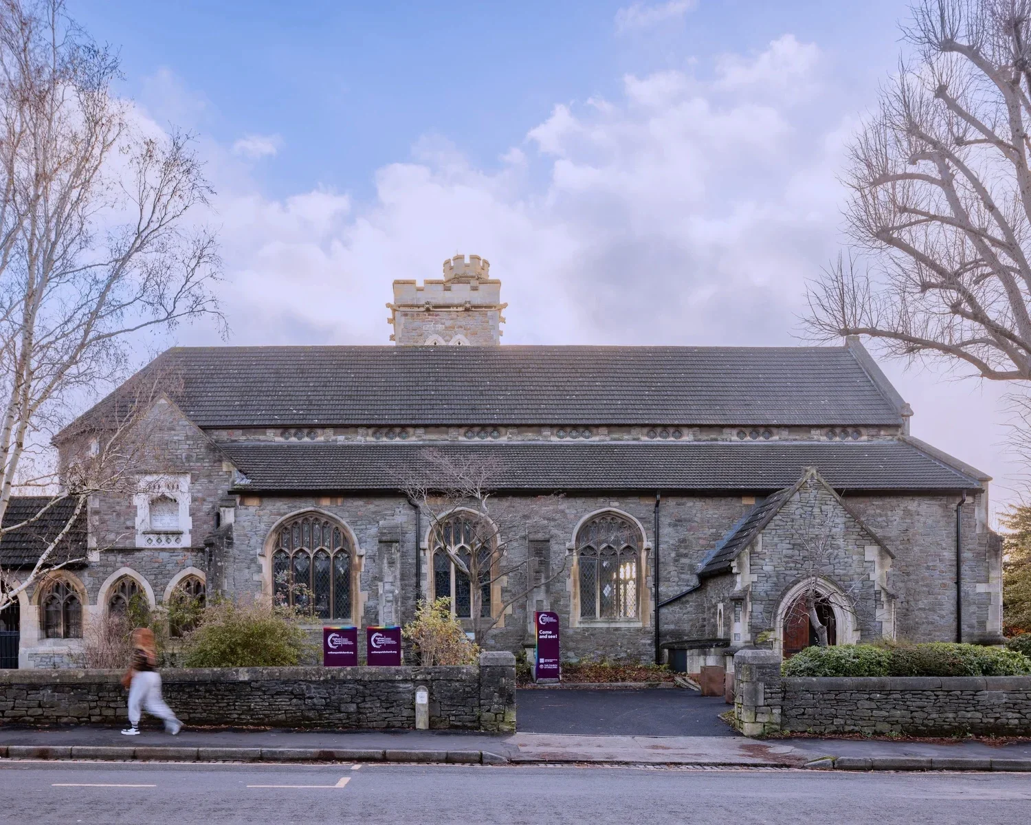 Stone church with arched stained glass windows, a tiled roof, and a small tower against a partly cloudy sky; trees and a pedestrian on the sidewalk in front.
