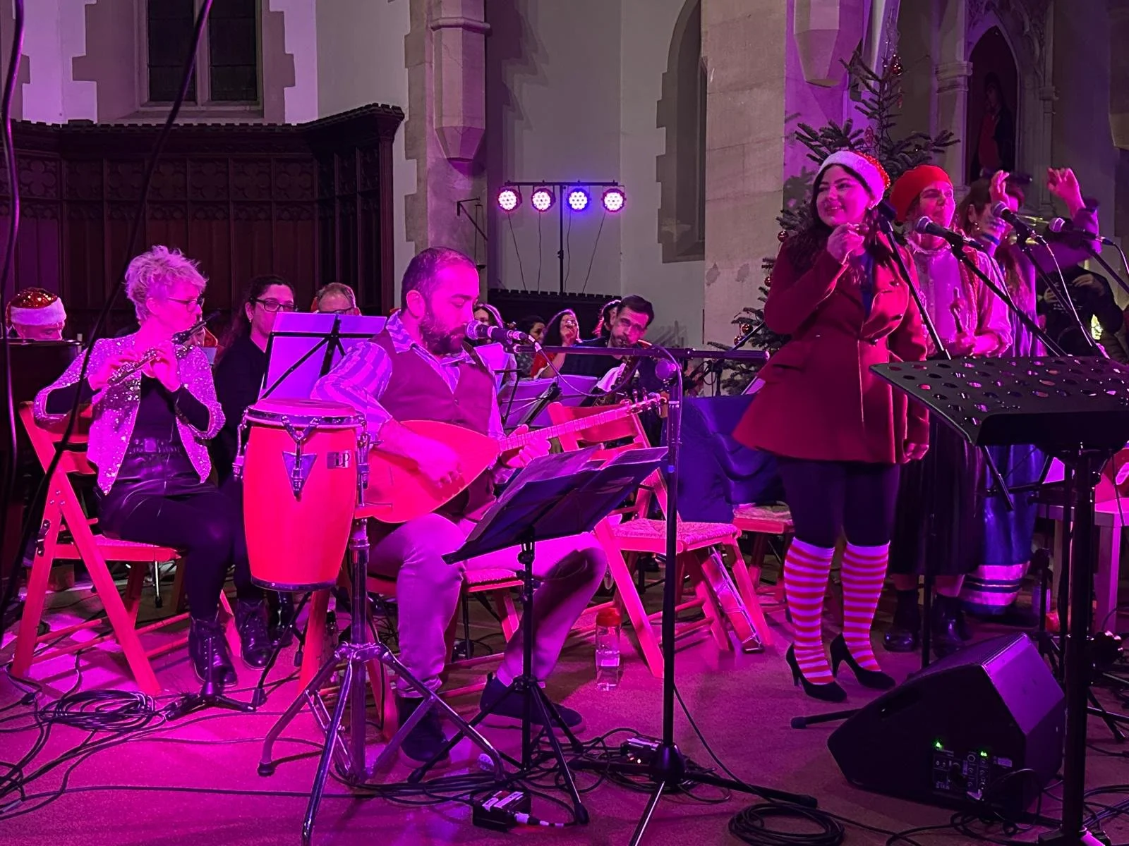 A Christmas music performance inside a decorated church with Christmas trees, featuring a group of musicians and singers. The performers are dressed in festive outfits, with some wearing Santa hats and cheery attire, illuminated by colorful stage lights.