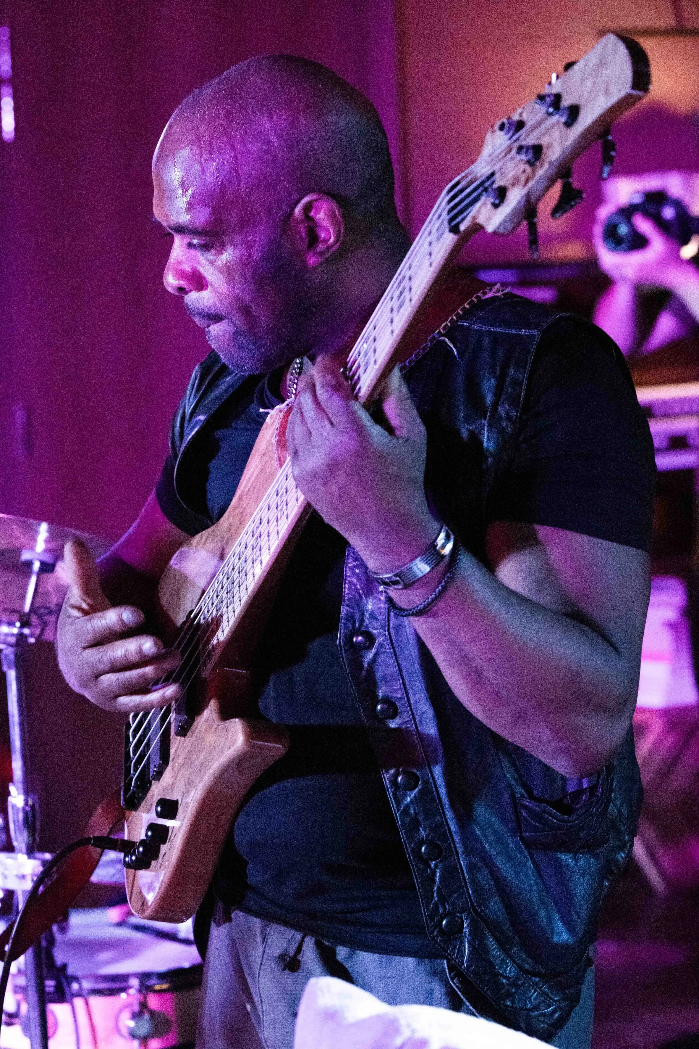 A man with a shaved head playing an electric guitar in a dimly lit room with pink and purple lighting.