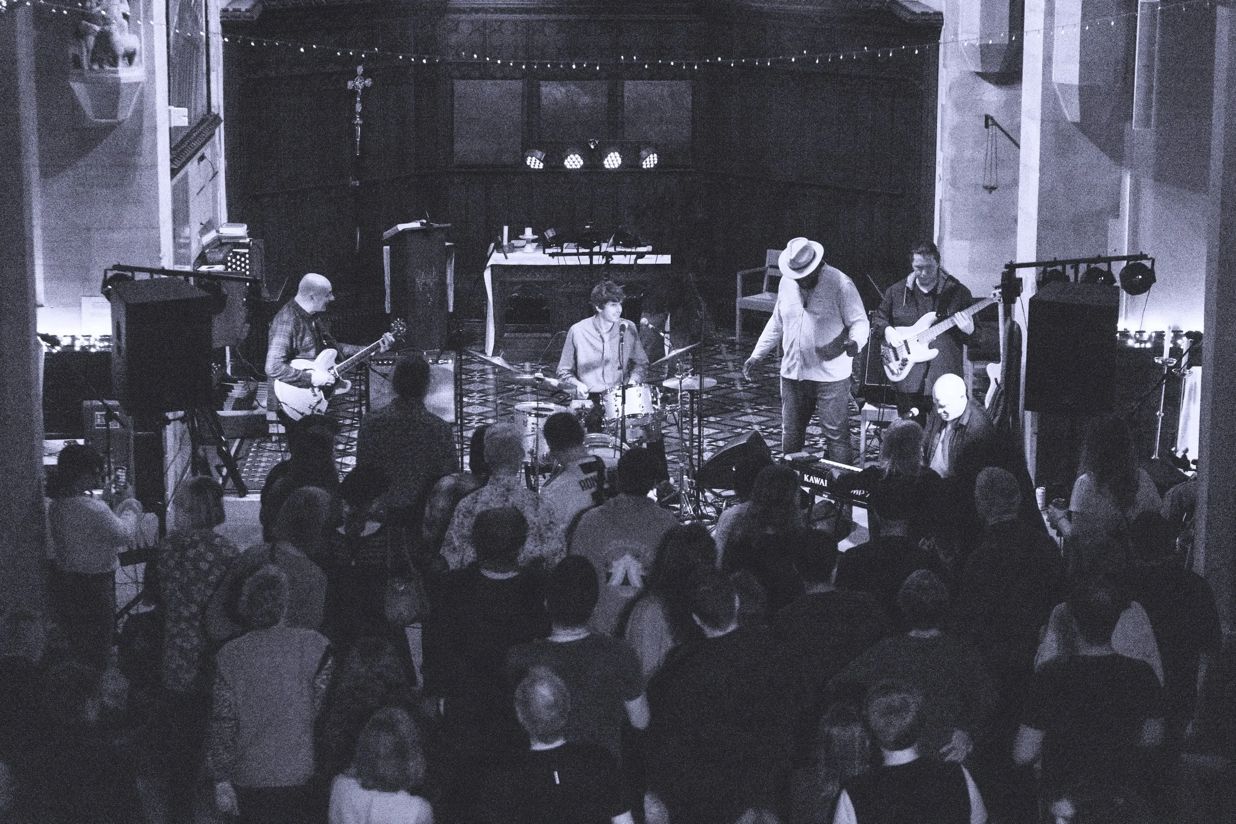 A band performs on stage in a dimly lit venue with an audience watching. The band includes a guitarist, a drummer, a bassist, and a vocalist with a hat. The stage features microphones, speakers, and musical equipment with an audience in foreground.