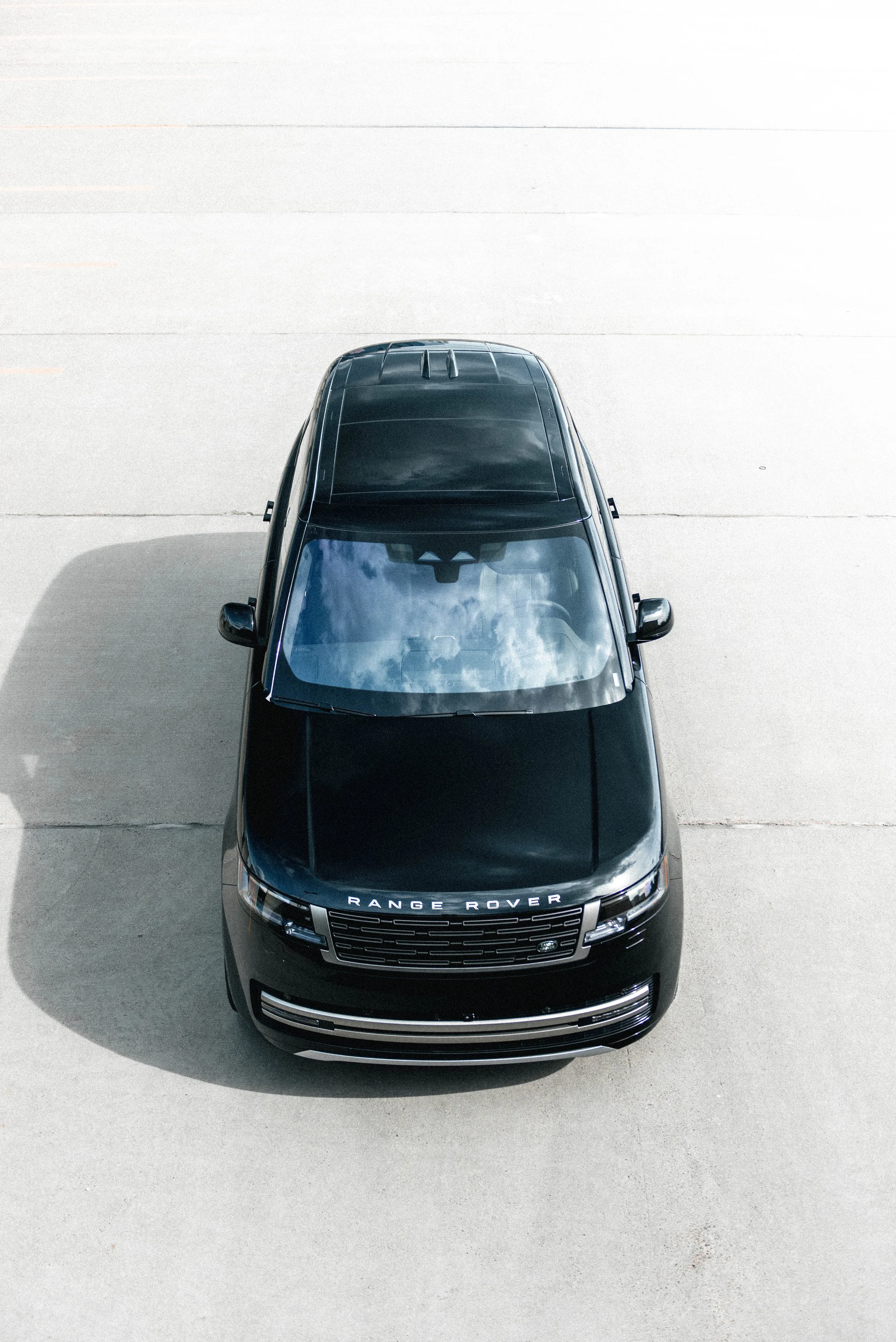 A black Range Rover SUV parked on a concrete surface, viewed from above, with the sun casting shadows and reflections on the roof.