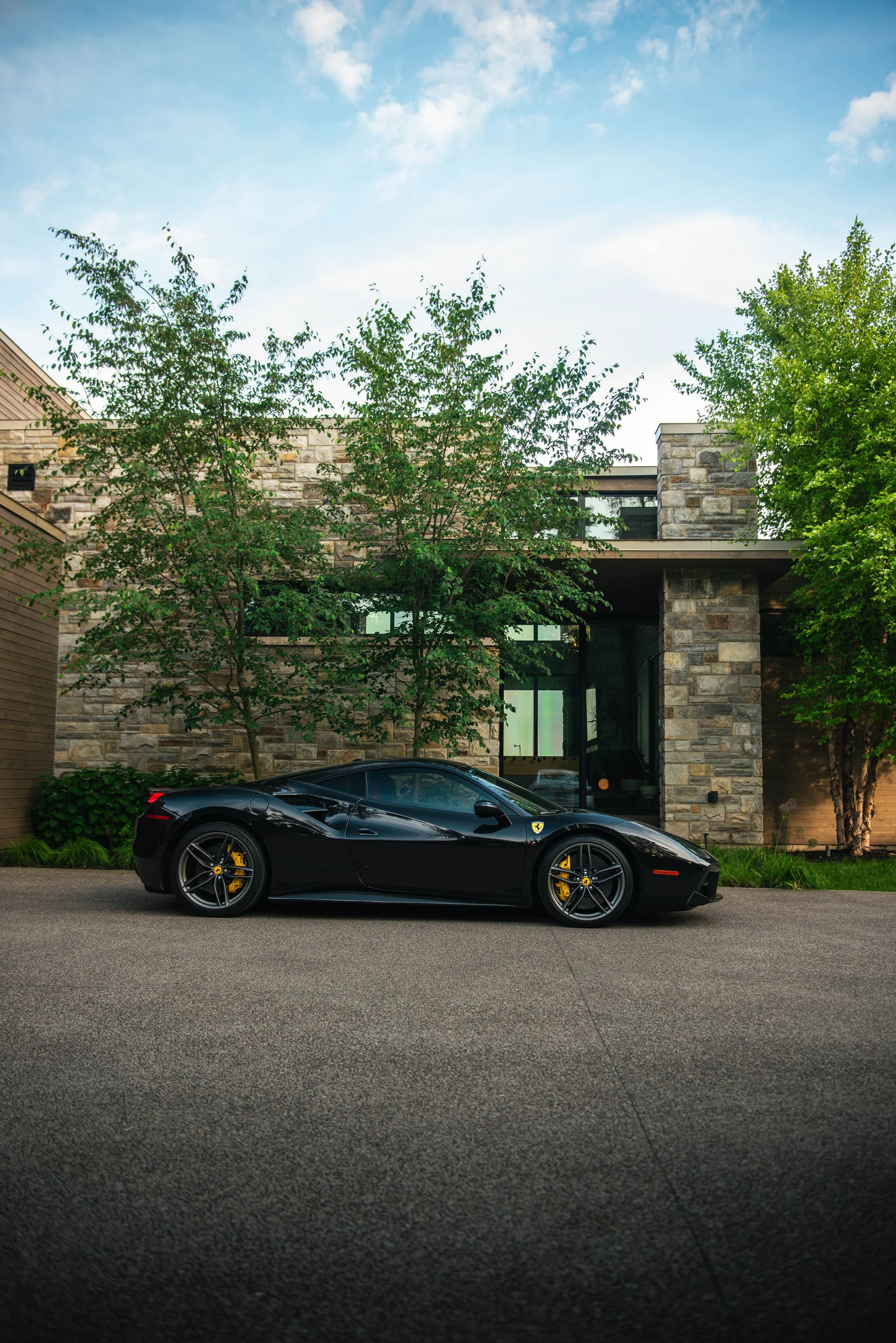 A black sports car parked in front of a modern house with stone exterior and large windows, surrounded by trees