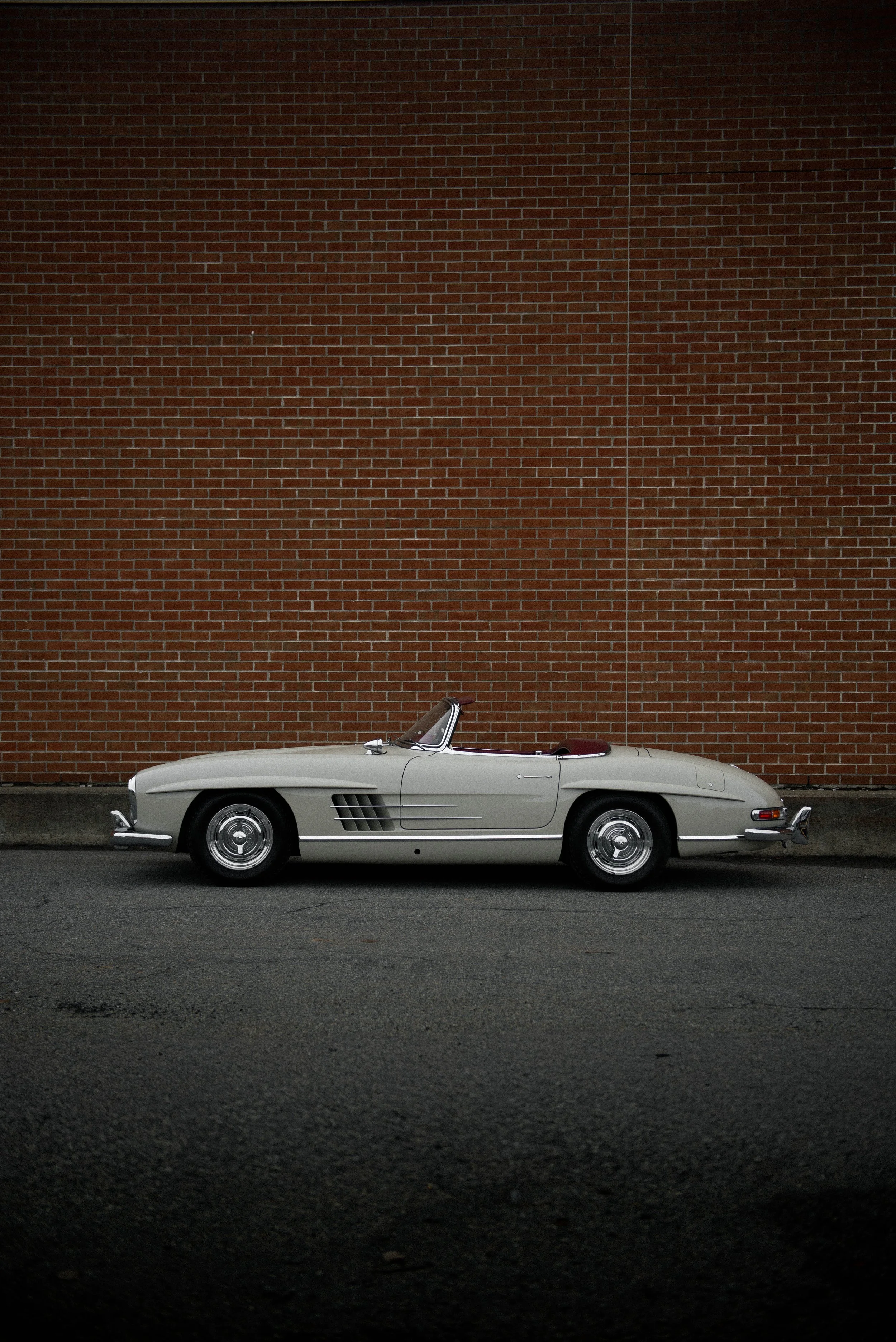 A white vintage convertible car parked on dark asphalt in front of a red brick wall.