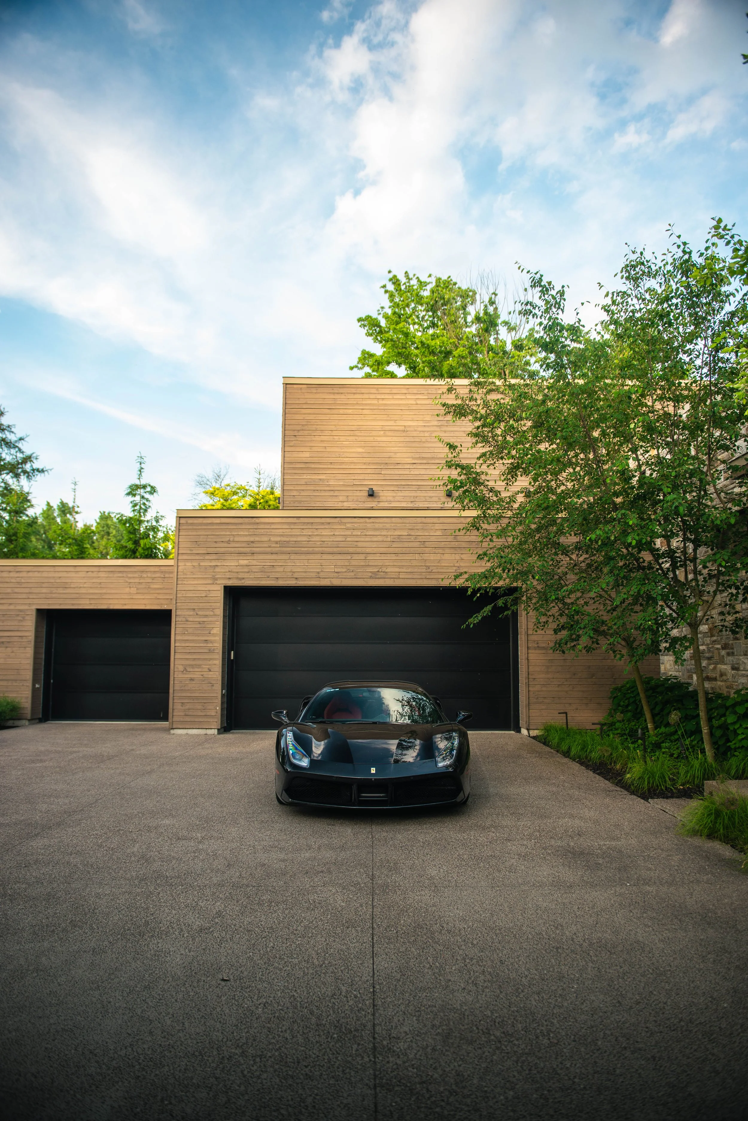 Black sports car parked in front of a modern house with a wooden facade, surrounded by greenery and trees, under a partly cloudy sky.