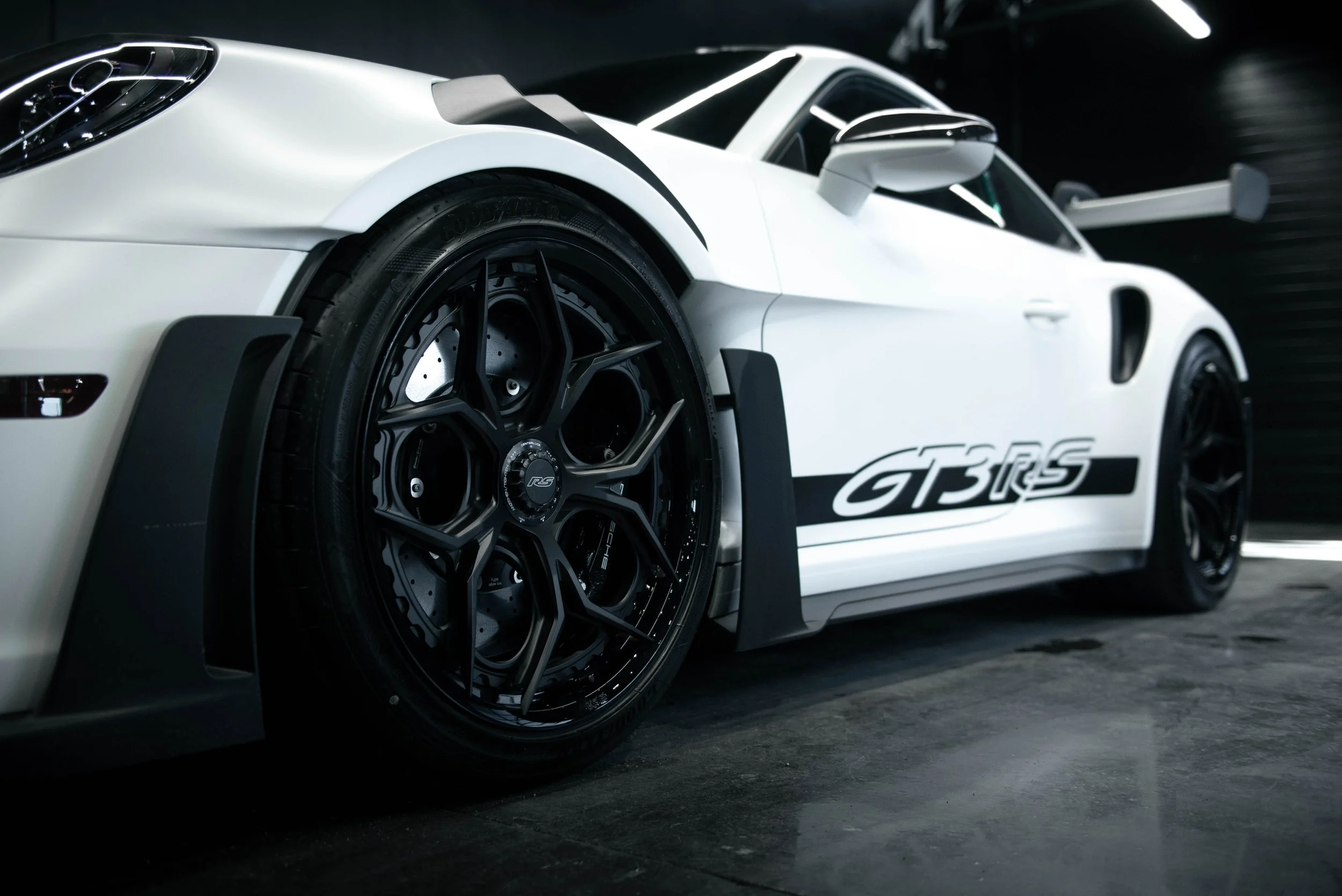 Close-up of a white race car with black wheels and GT3 RS decals, parked in a low-light garage.