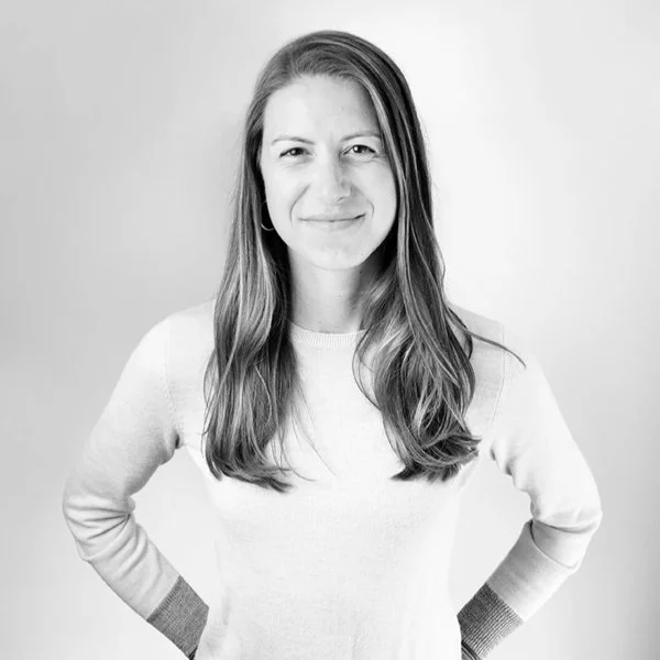 Black and white photo of a woman with long hair, smiling with one eye slightly squinted, wearing a light-colored sweater with darker cuffs, standing against a plain background.