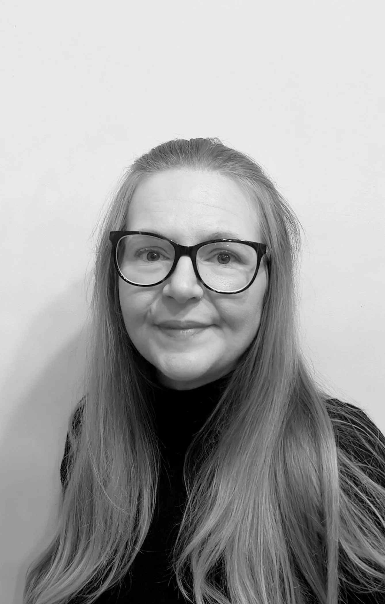 Black and white photo of a woman with long hair, wearing glasses, smiling in front of a plain wall.