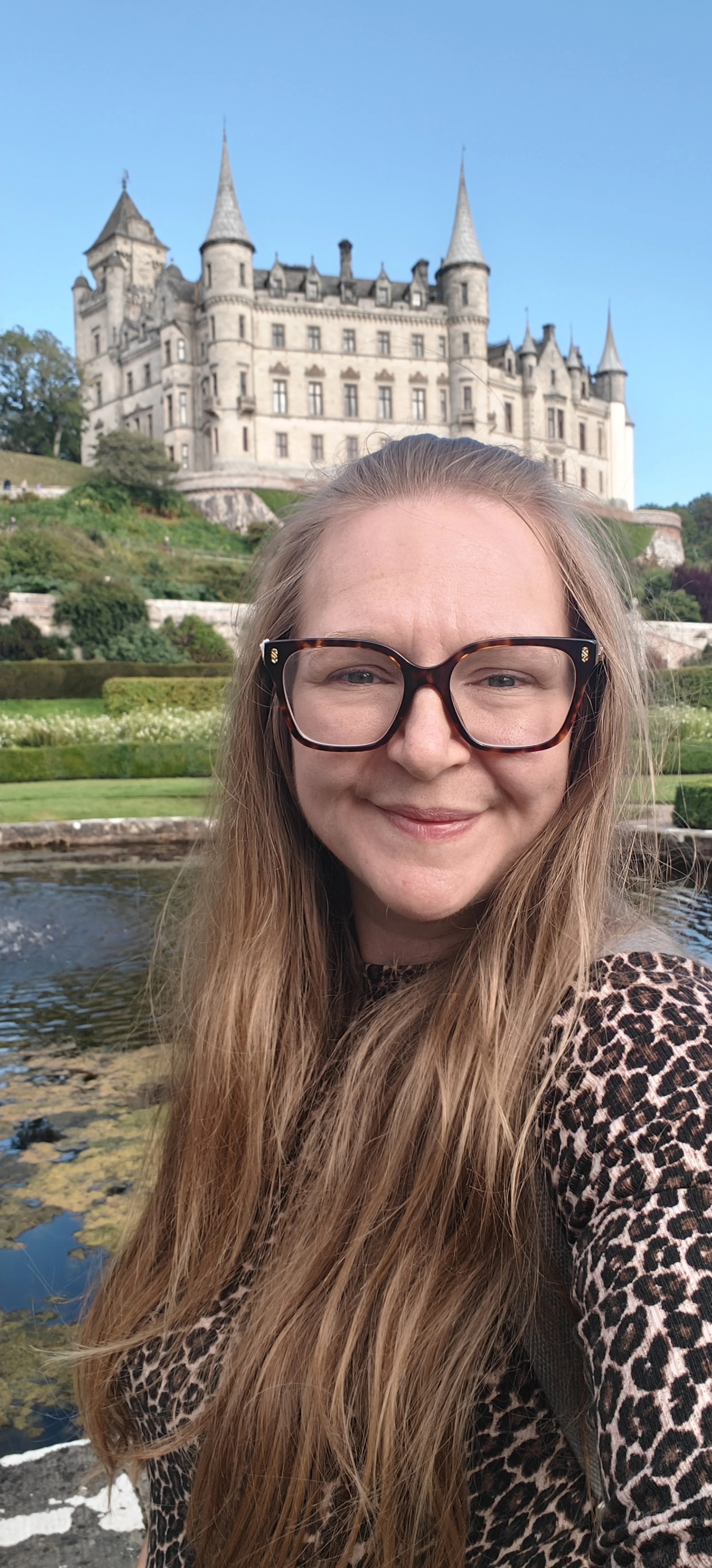A woman with long, wavy hair, wearing glasses and a leopard print top, is smiling while taking a selfie in front of a castle with turrets and spires, surrounded by greenery and a pond.