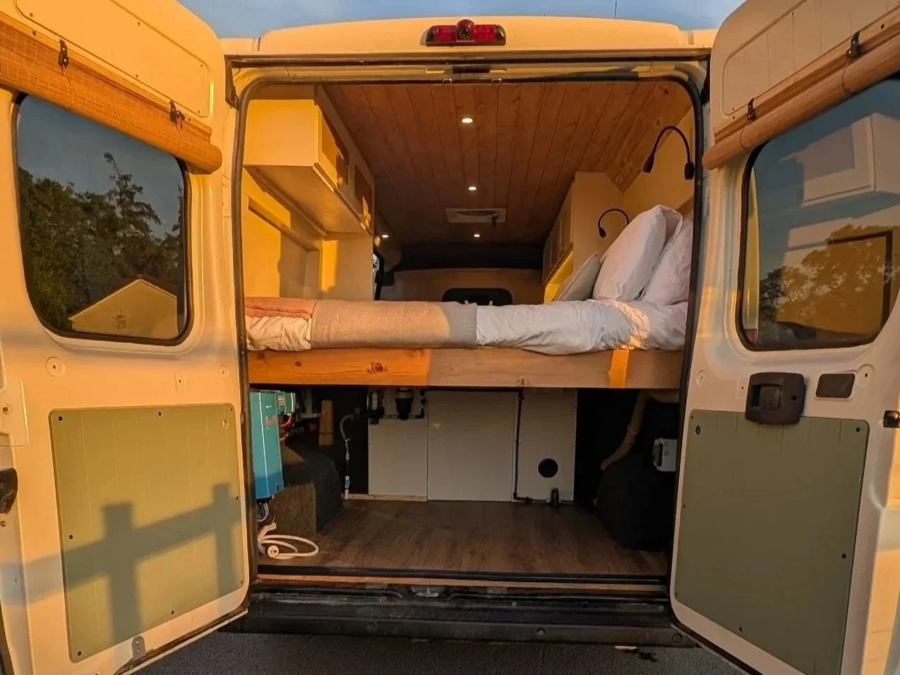 View of a bespoke camper van interior with a bed, wooden ceiling, and side windows reflecting trees and a house outside.