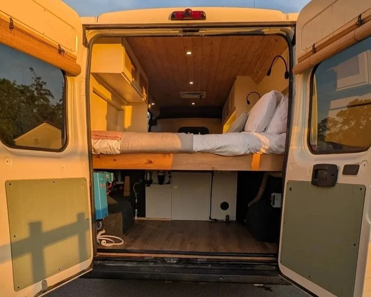 Interior of a camper van with a raised bed made of wood, white bedding, and pillows, with wooden panel ceiling and small built-in shelves, illuminated by ceiling lights, with windows on either side showing outdoor scenery.