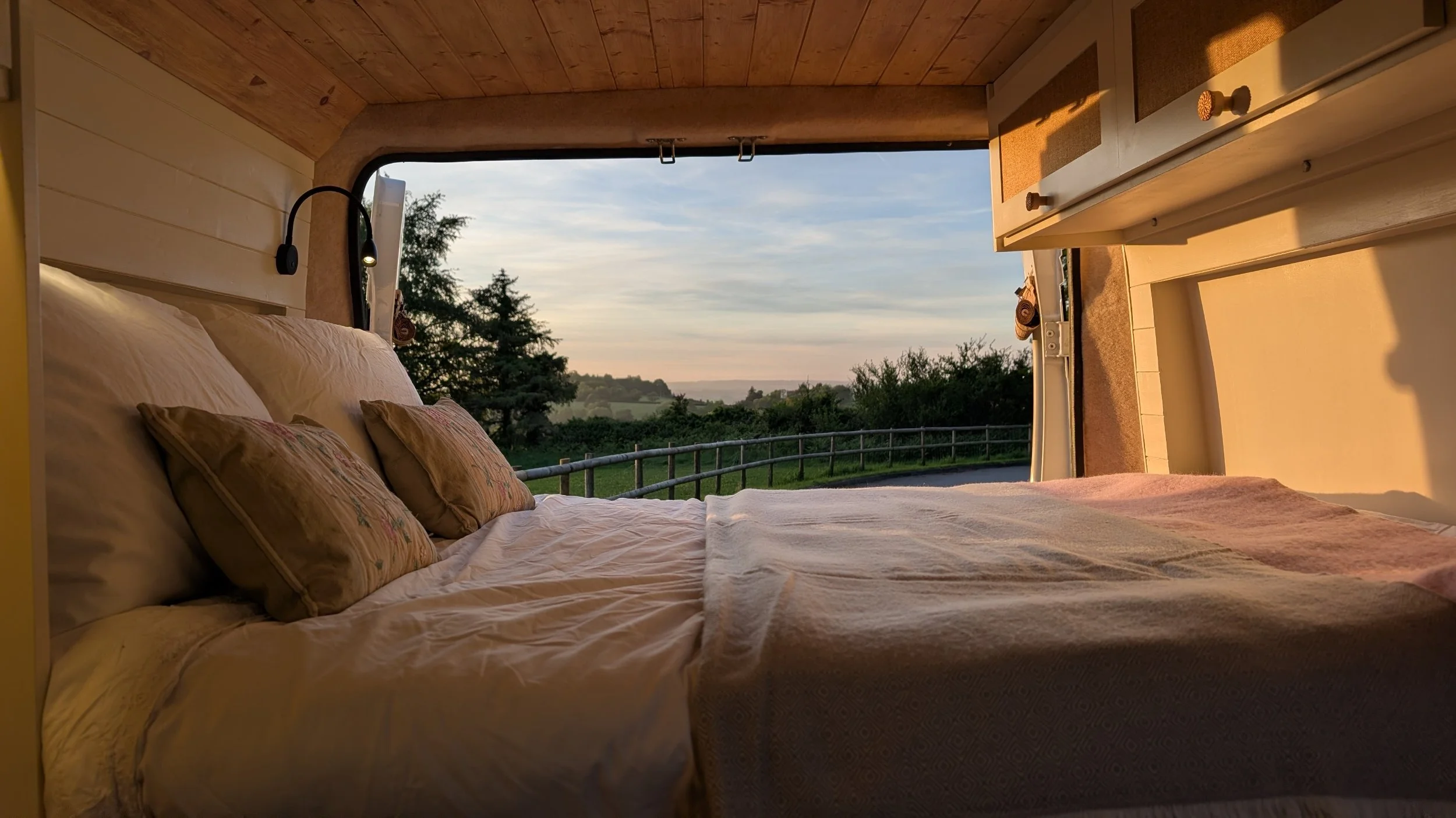 A cozy Campervan bedroom with a large bed, fluffy pillows, and a view of trees and a landscape through a large window at sunset.