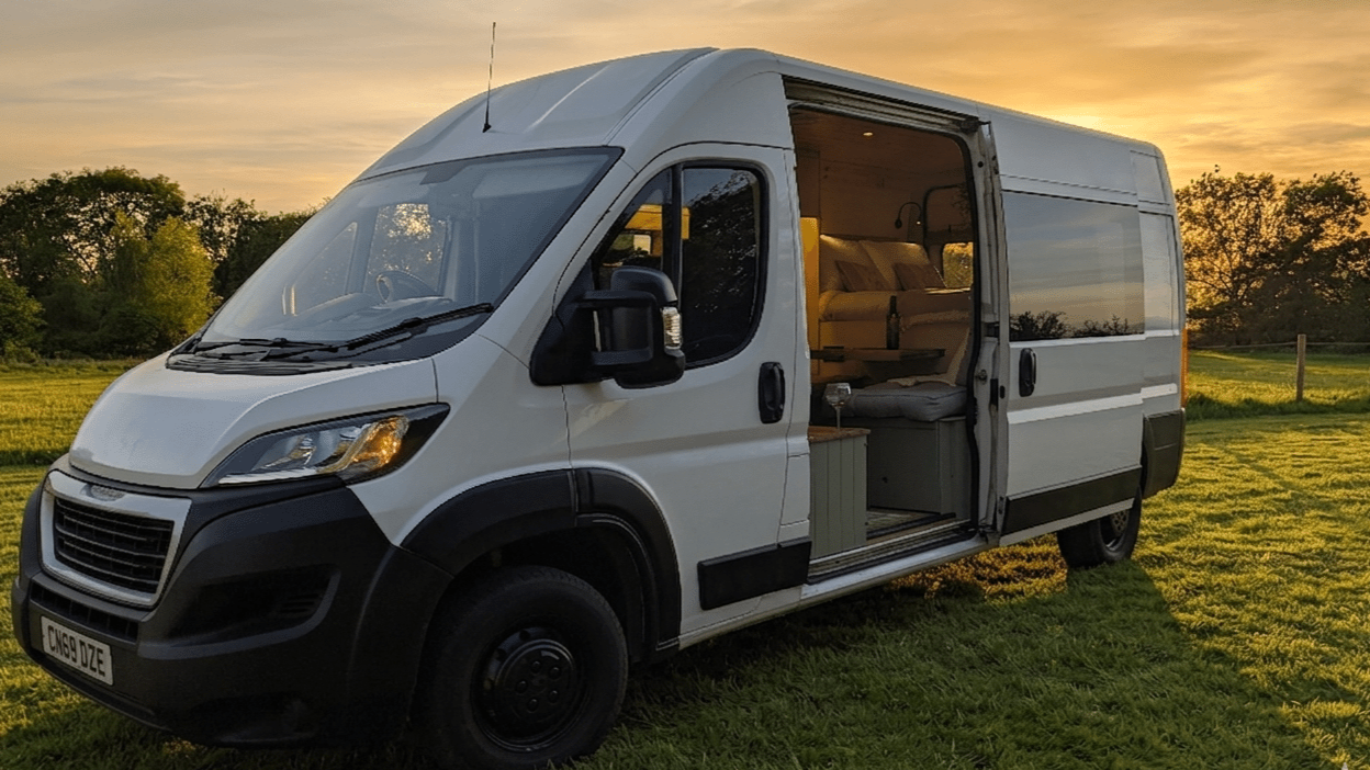 White handcrafted, bespoke, camper van with open sliding door, parked on a grassy field during sunset, showing a cozy interior with seating and a small table.