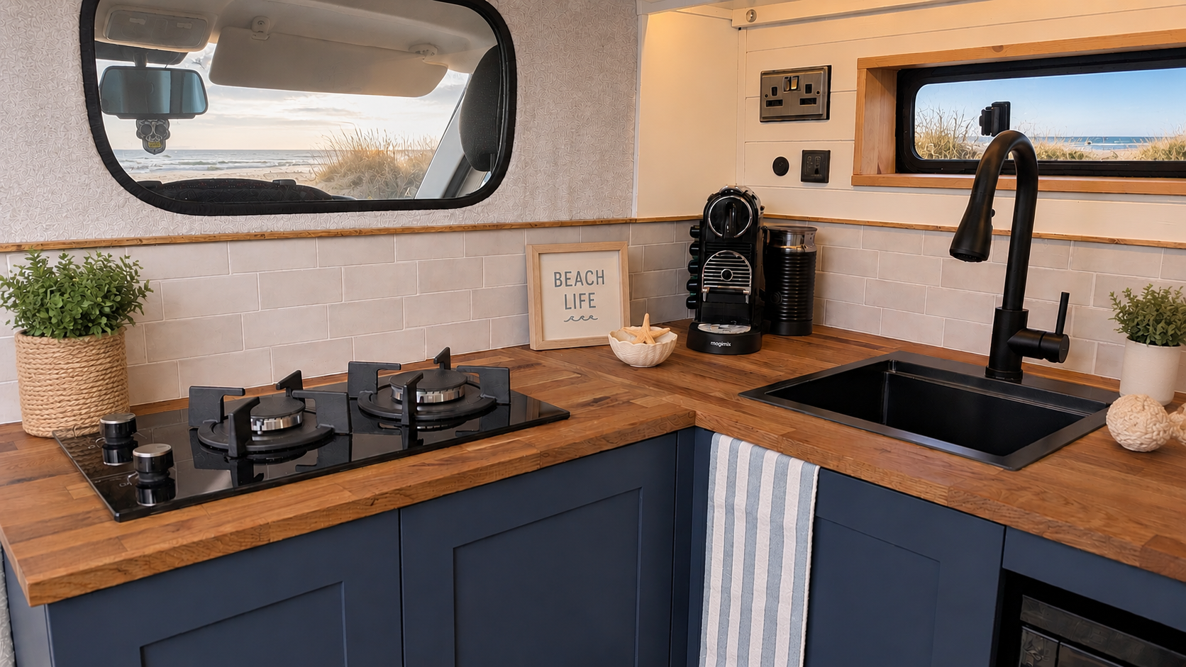 Cozy kitchen with wooden countertops, navy cabinets, black sink, coffee maker, potted plants, beach-themed decor, and windows showing beach scenery outside.
