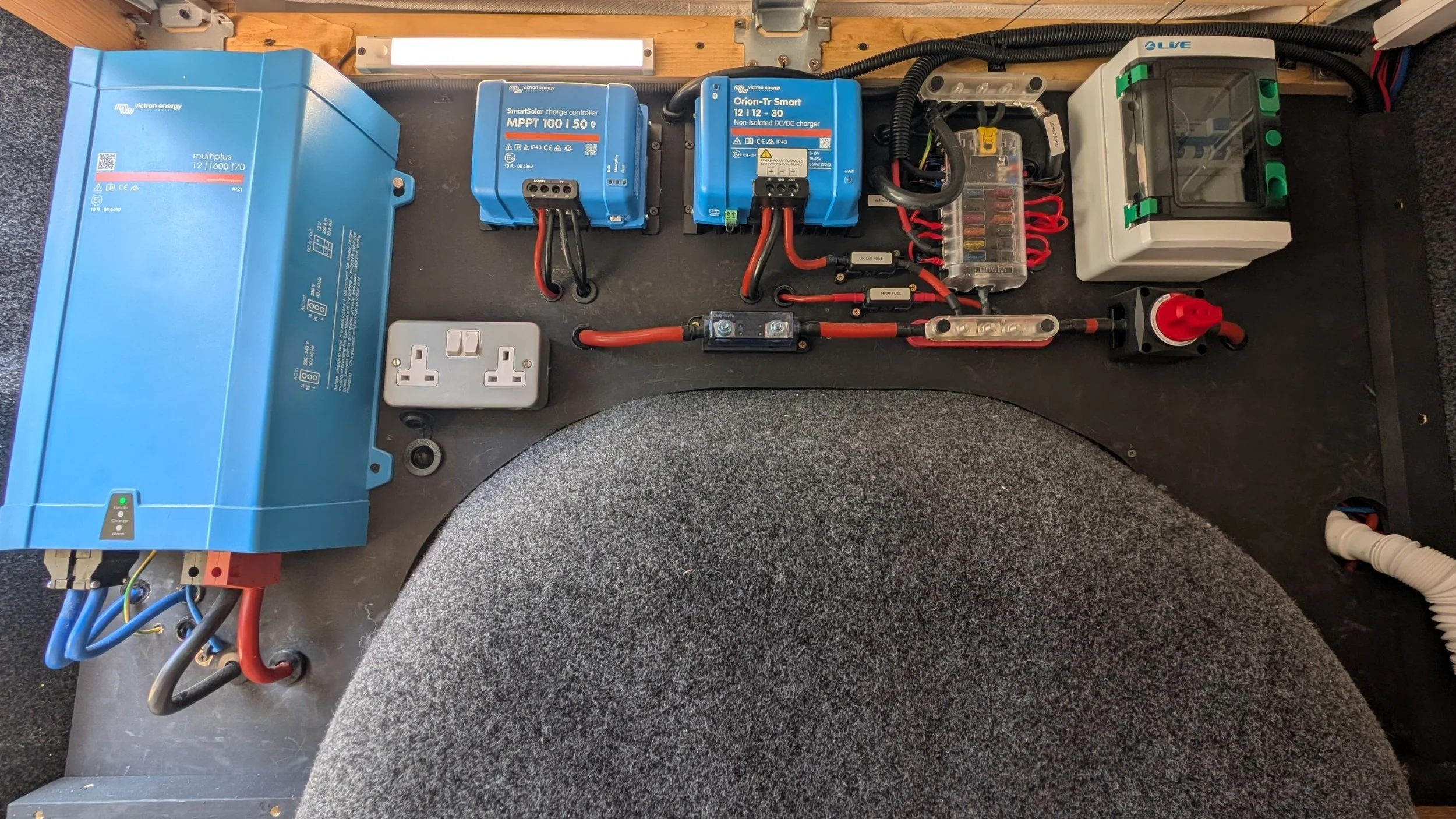 Image showing a collection of electrical and electronic components mounted on a black surface. The components include a large blue inverter, two blue charge controllers labeled 'MPPT 100 | 50' and 'Orion-Tr Smart 12 | 12 - 30,' a plastic fuse box with red wires, a small gray power outlet, and a small red and black plug. There are various wires and cables connected to the devices.