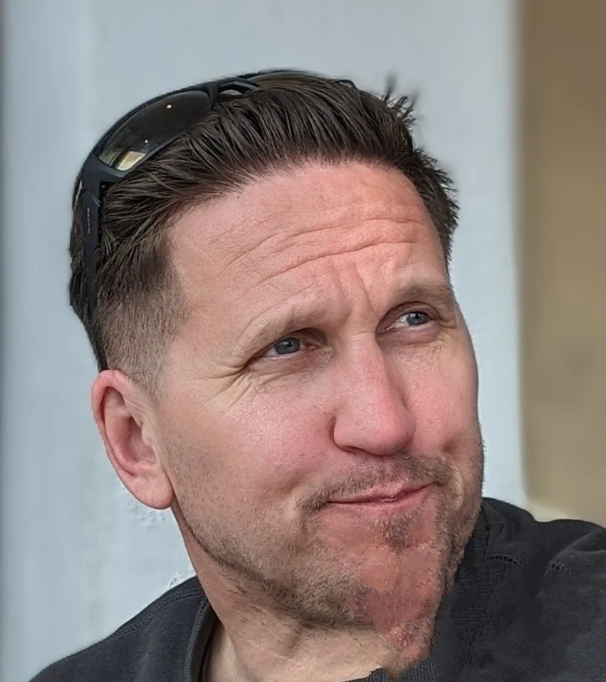 Close-up of a man with short brown hair, blue eyes, and a slight beard, wearing sunglasses on his head, looking to the side with a slight smile.