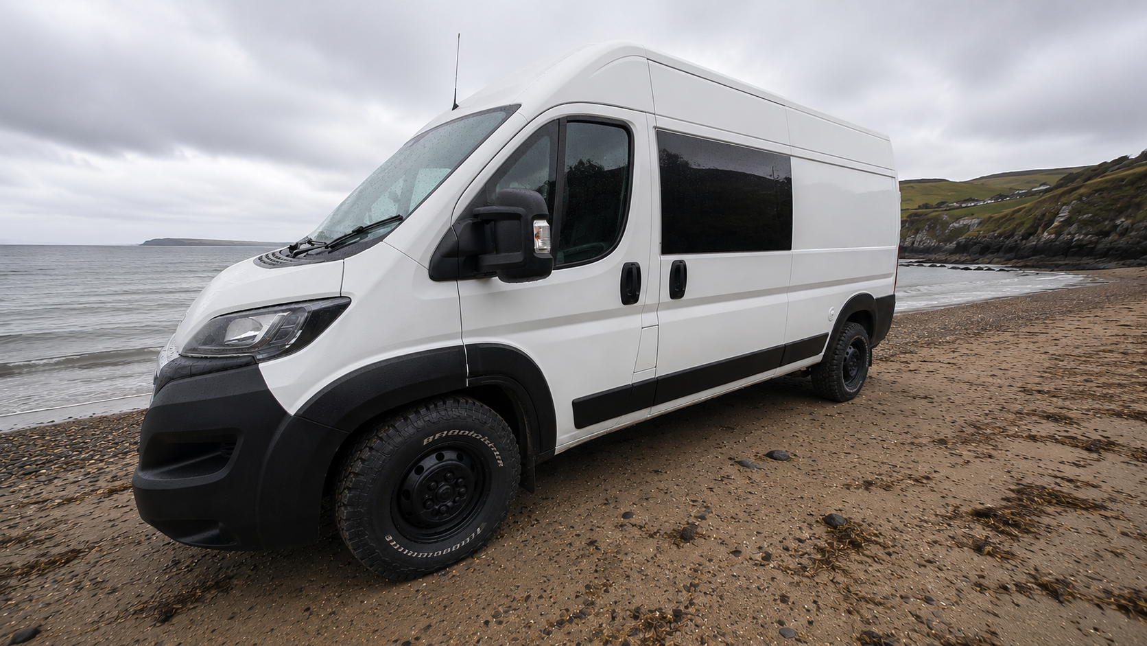 White cargo van parked on a sandy beach near the ocean with grassy hills and cliffs in the background under cloudy skies.