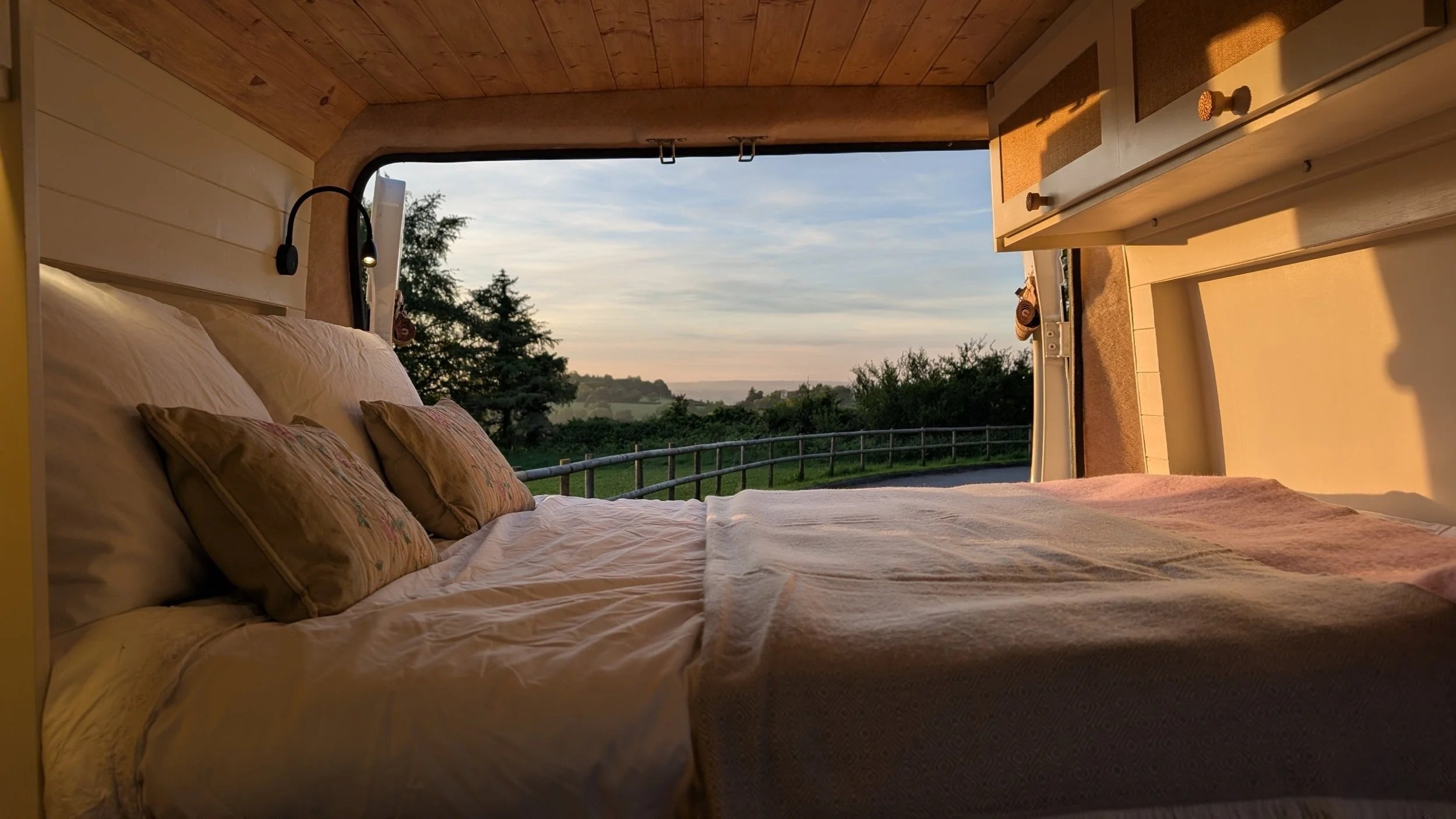 A cozy bedroom with a bed made with light-colored bed linens and floral pillows. The room has a large open window showing a scenic countryside view with trees and a fence under a partly cloudy sky during sunset.
