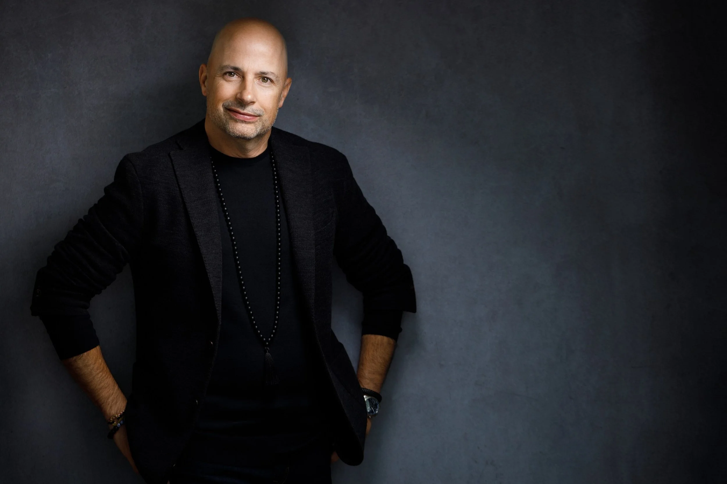 A portrait of a middle-aged man with a shaved head and a slight beard, wearing a black jacket and a black shirt, standing against a dark gray background.