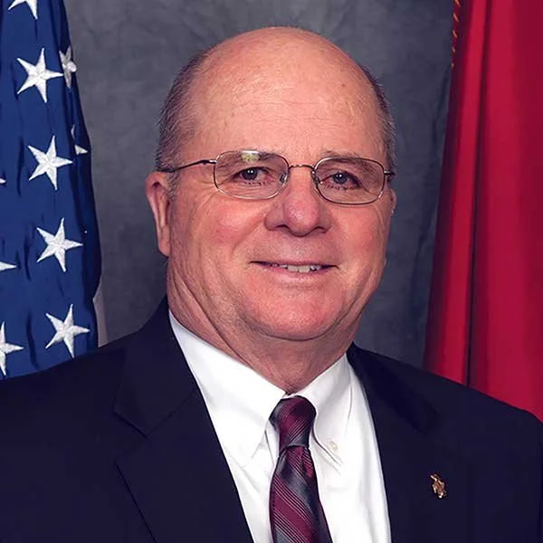 A middle-aged man in a suit and tie, wearing glasses, smiling, standing in front of an American flag and another red flag.