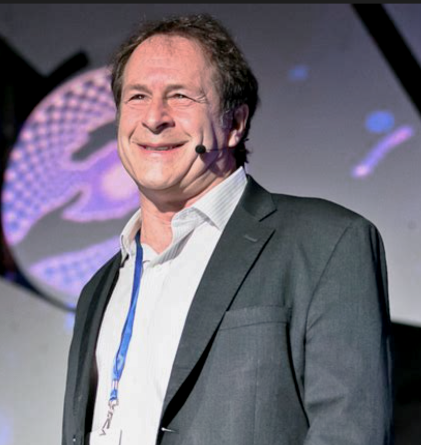A man in a suit smiling on stage with a headset microphone, standing in front of a screen with a digital graphic.