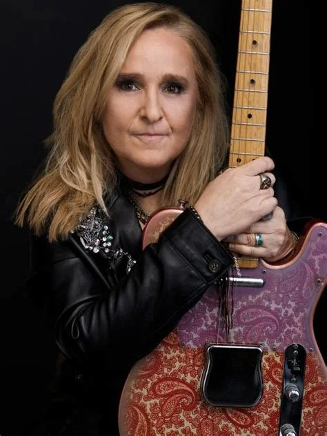 A woman with blonde hair holding an electric guitar, wearing a black leather jacket and rings, against a black background.