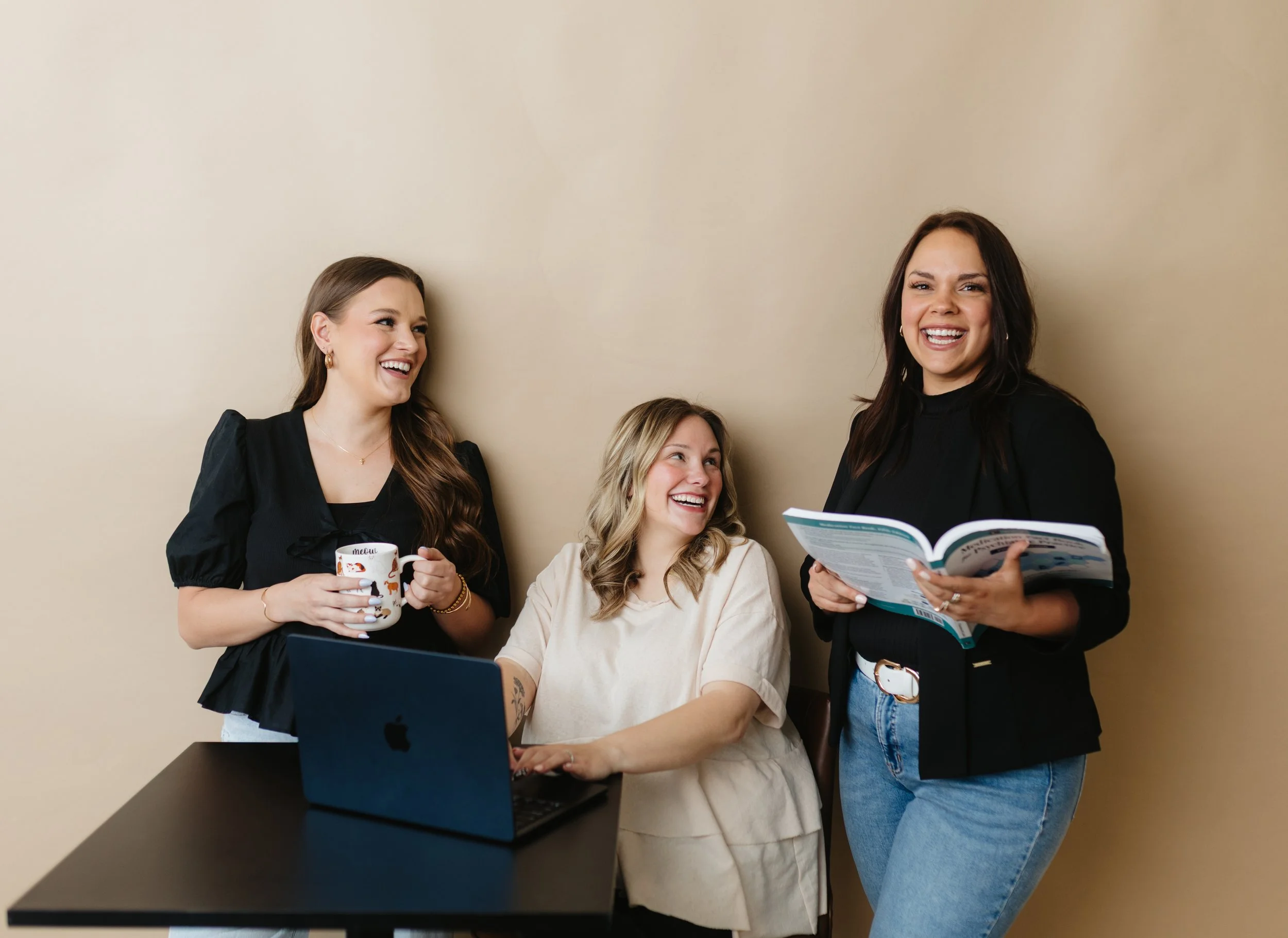 Four women gathered around a laptop, smiling and talking. One woman holds a mug, another holds a magazine, and the third appears to be looking at the magazine. They are in an indoor setting with a beige wall background.