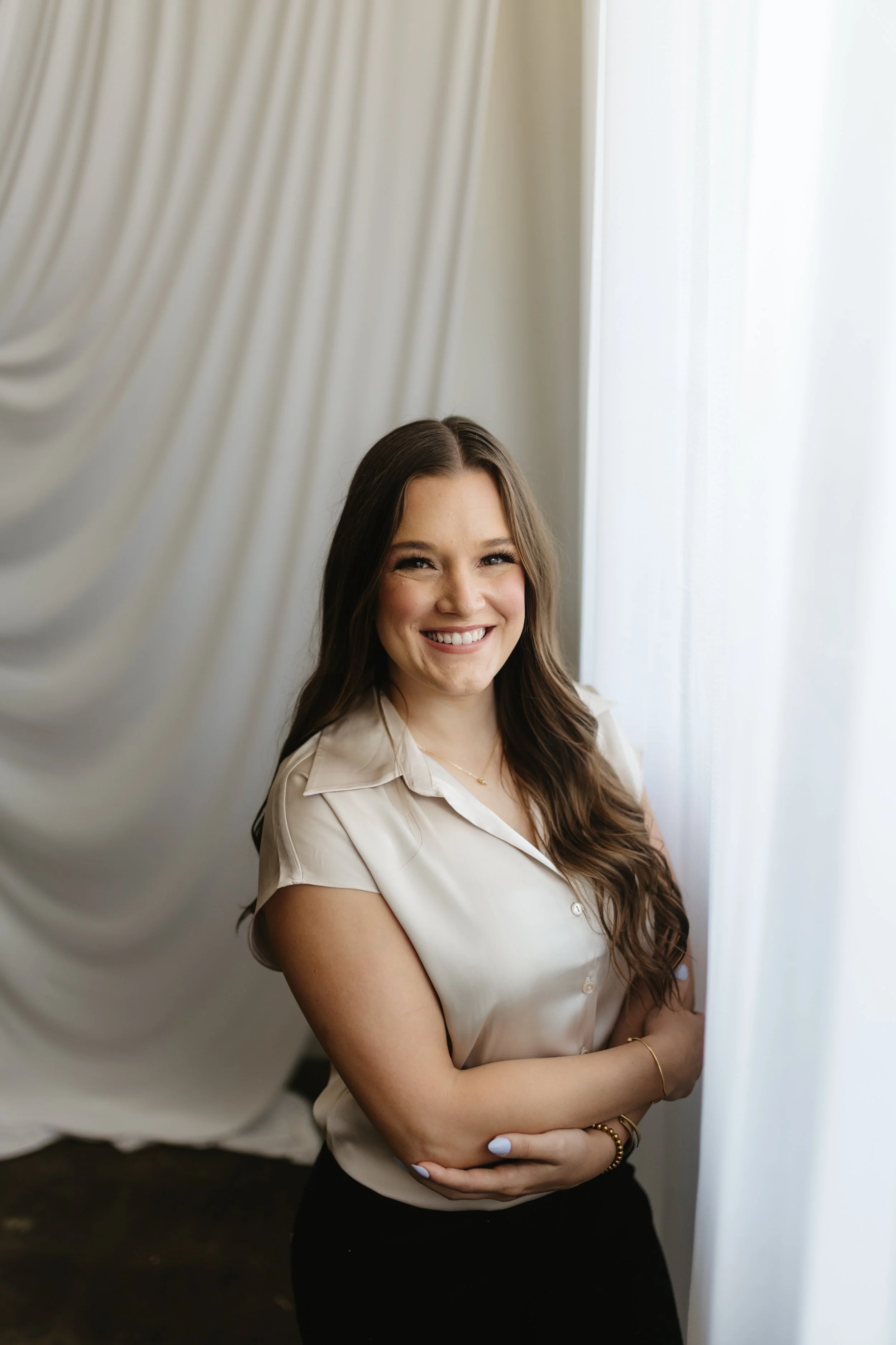 A young woman with long brown hair standing indoors near white curtains, smiling and crossing her arms.
