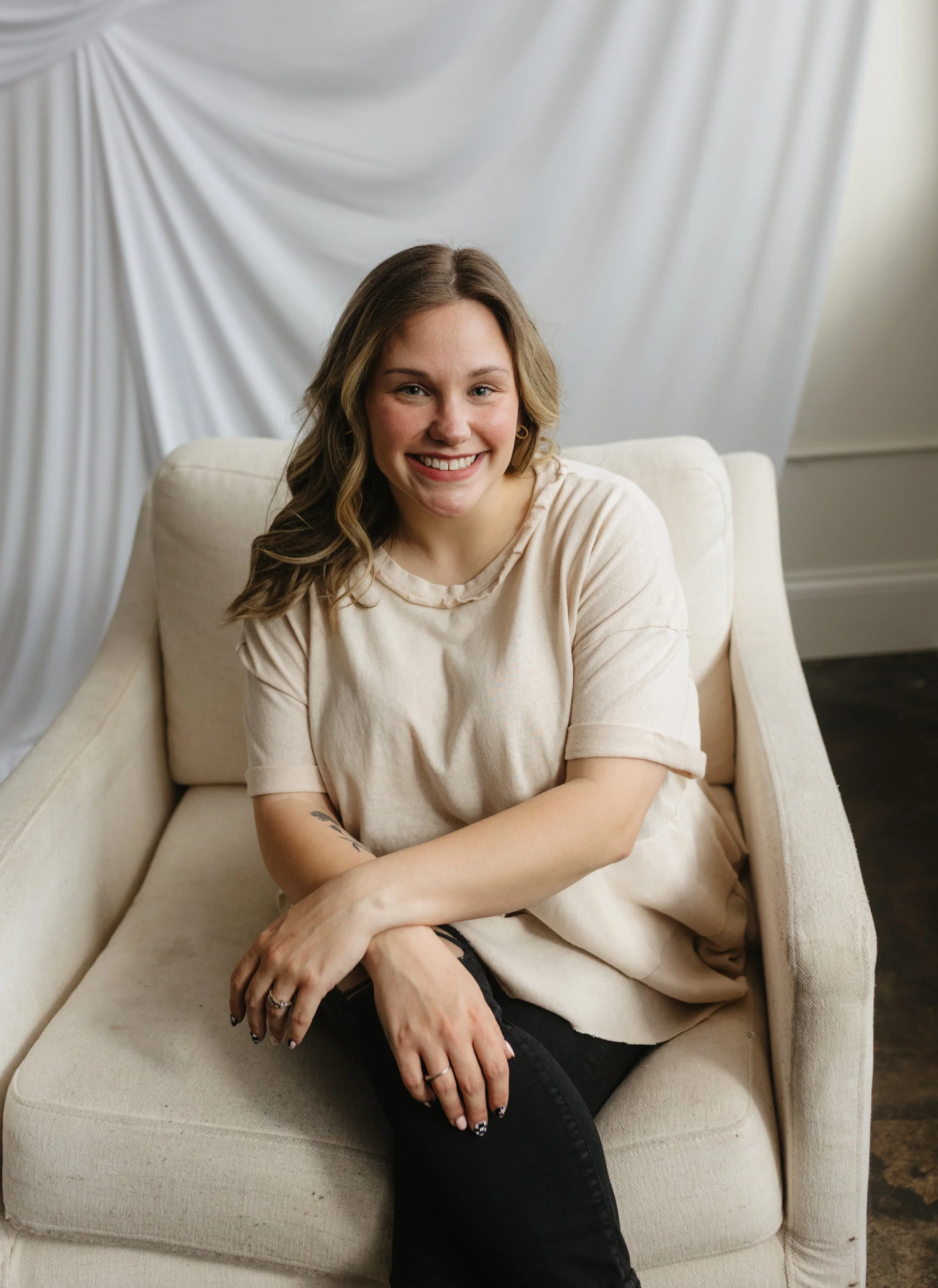 A young woman with shoulder-length wavy hair, smiling, sitting on a cream-colored sofa in front of a white draped backdrop, wearing a beige top and black ripped jeans.