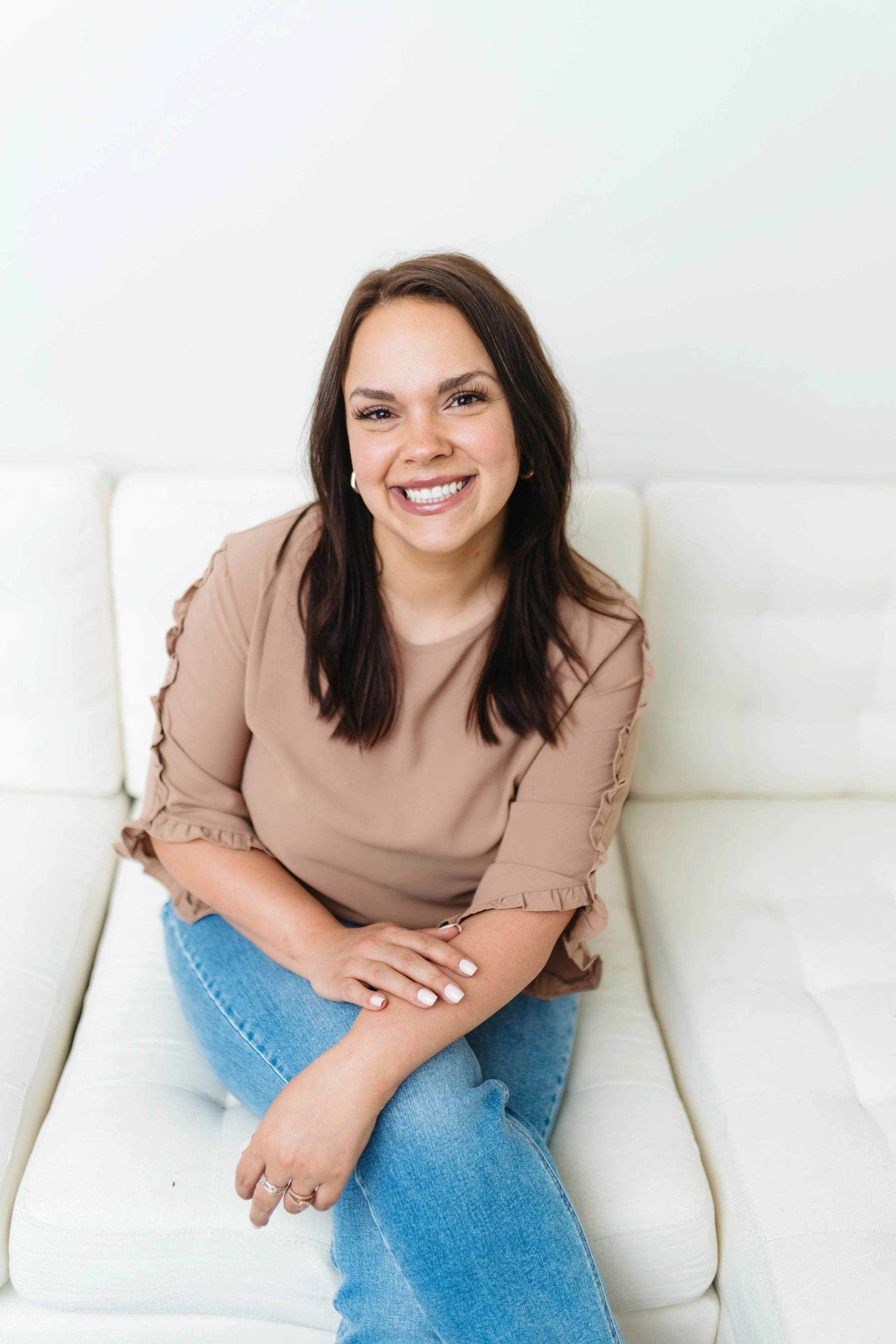 A smiling woman with dark brown hair, wearing a beige blouse and blue jeans, sitting on a white sofa in front of a plain white wall.