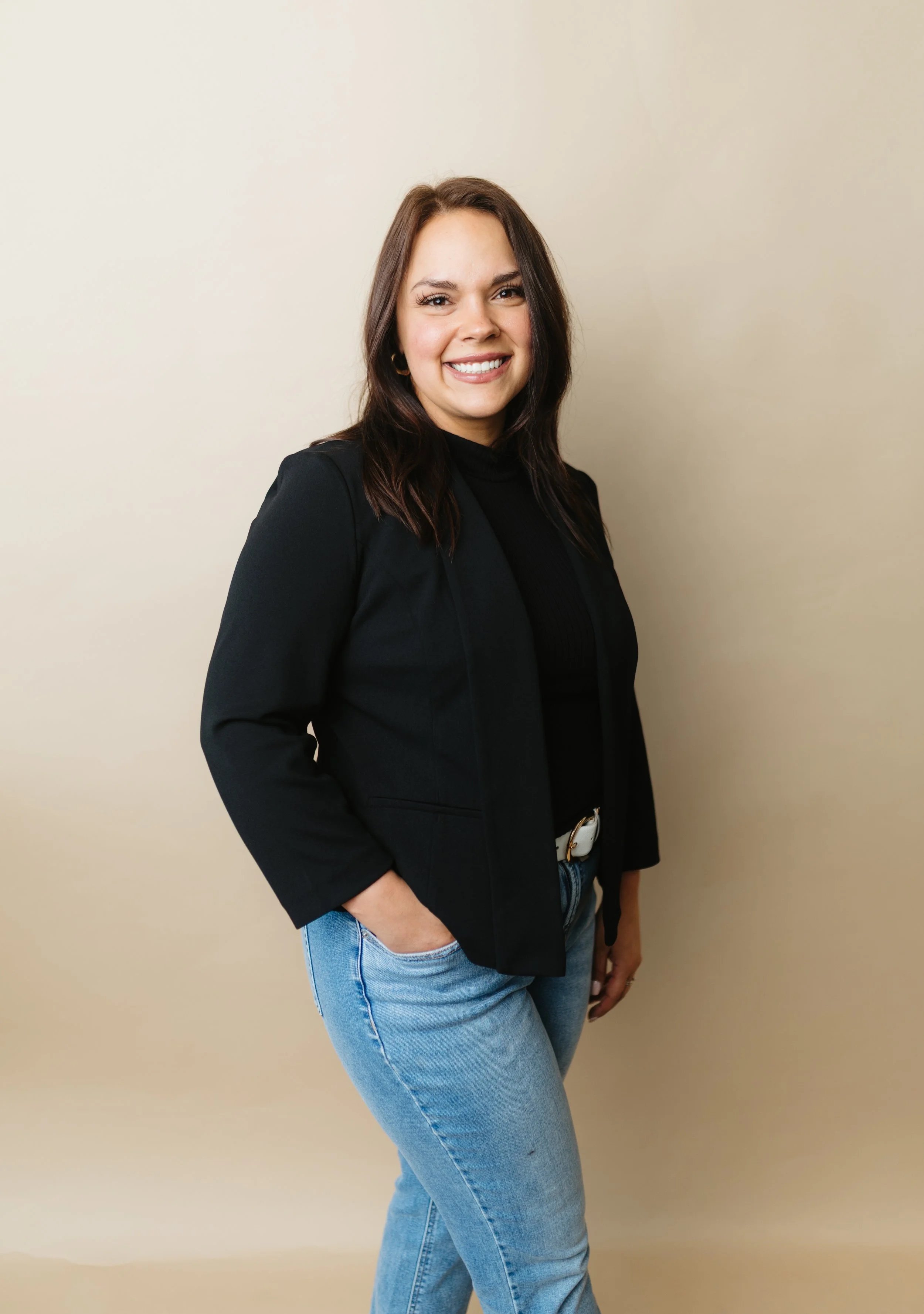 A woman smiling, wearing a black blazer and light blue jeans, standing against a plain light-colored wall.