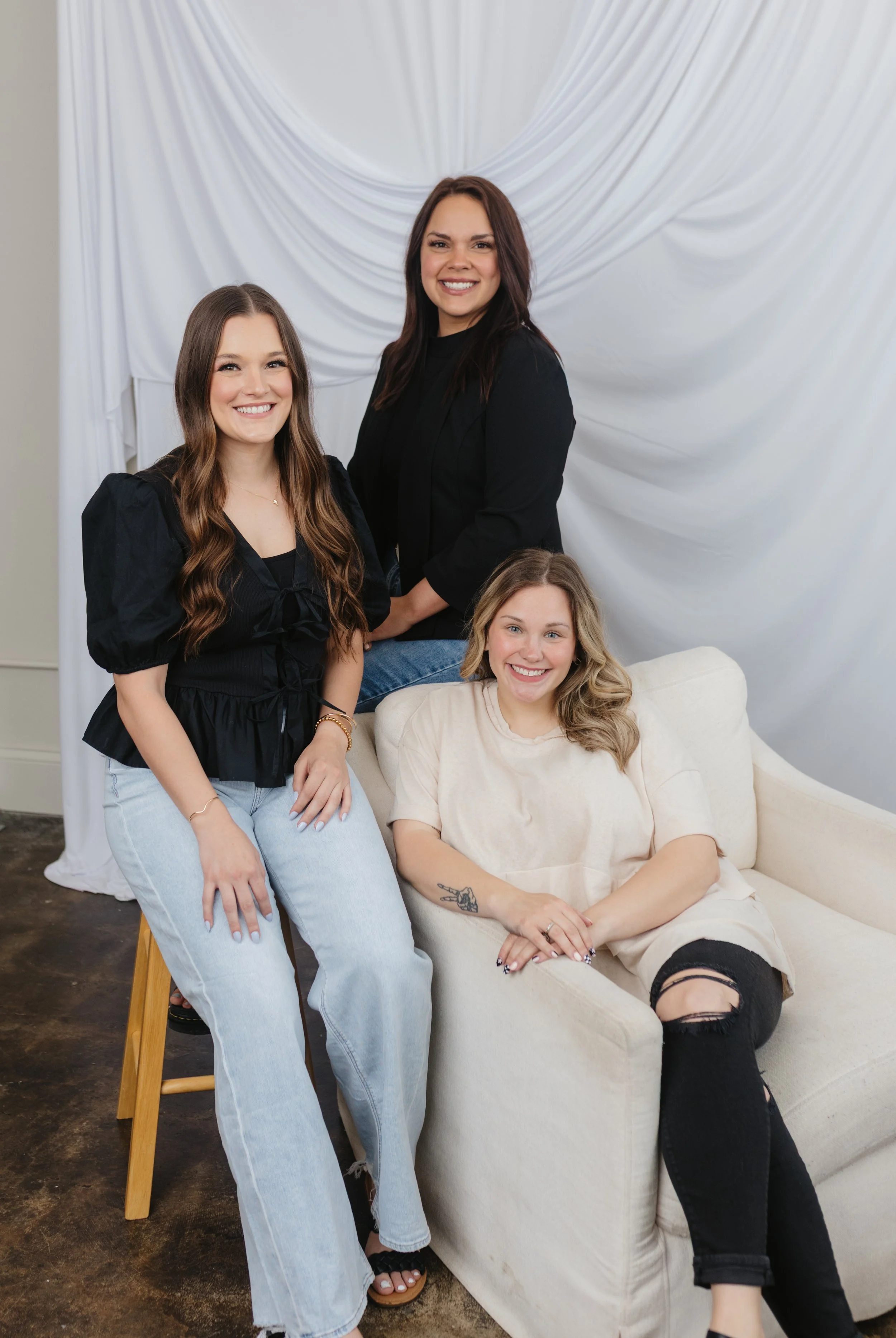Four women posing together in front of a white draped fabric backdrop; one seated on a cream-colored couch, others standing around her, all smiling.