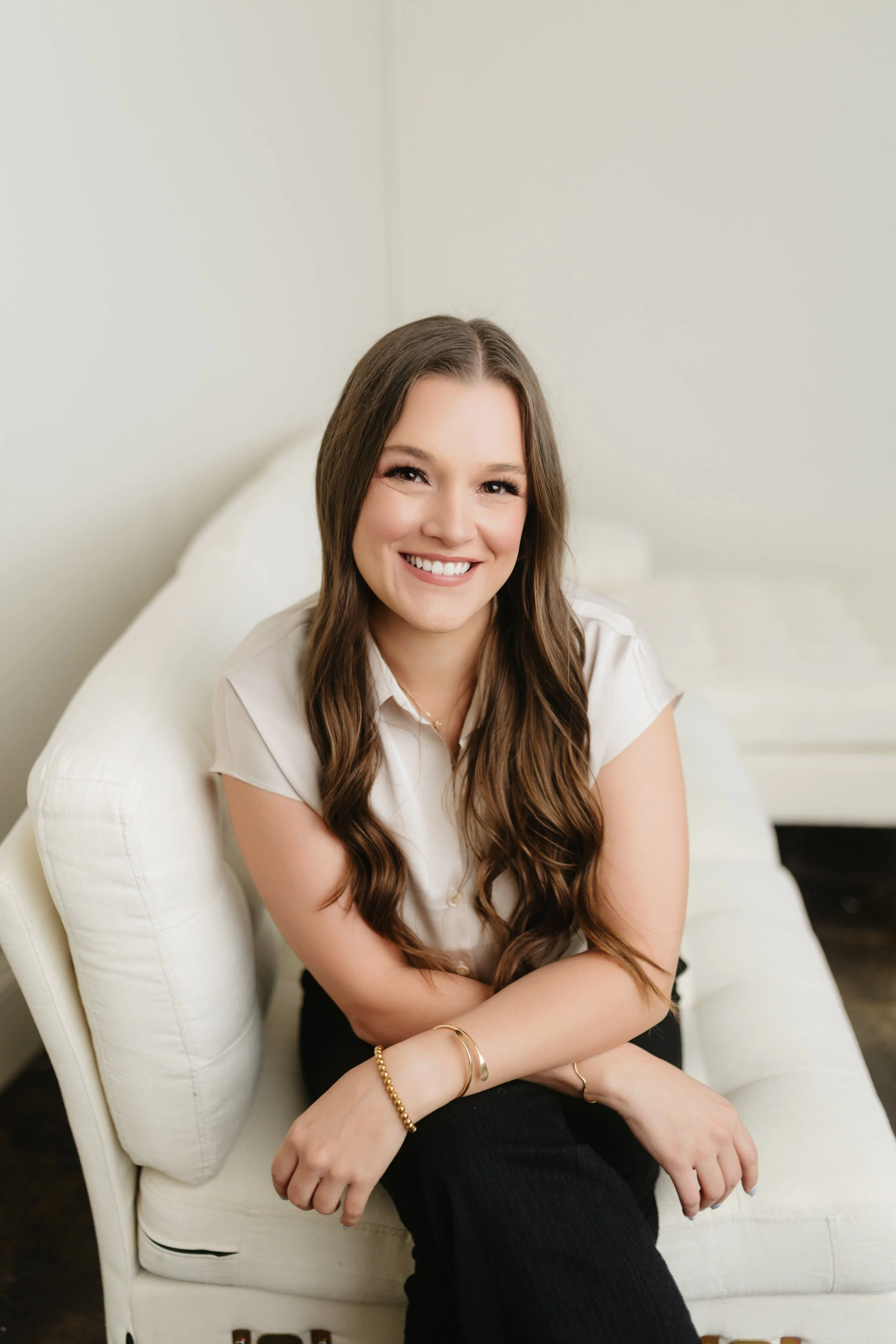 A young woman with long brown hair and a white blouse, smiling and sitting on a white armchair in a neutral-colored room.