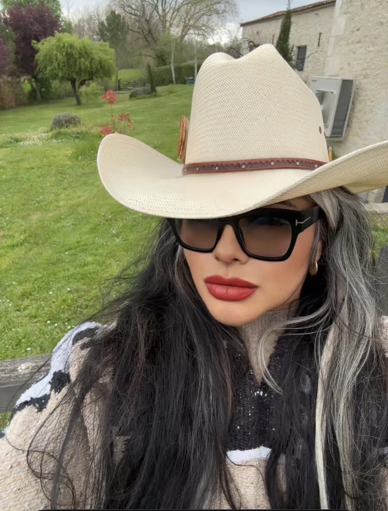 A woman with long dark hair, wearing black sunglasses, red lipstick, a beige wide-brimmed hat with a brown band, and a beige and black cow-patterned jacket, standing outdoors in a green grassy area with trees and a house in the background.