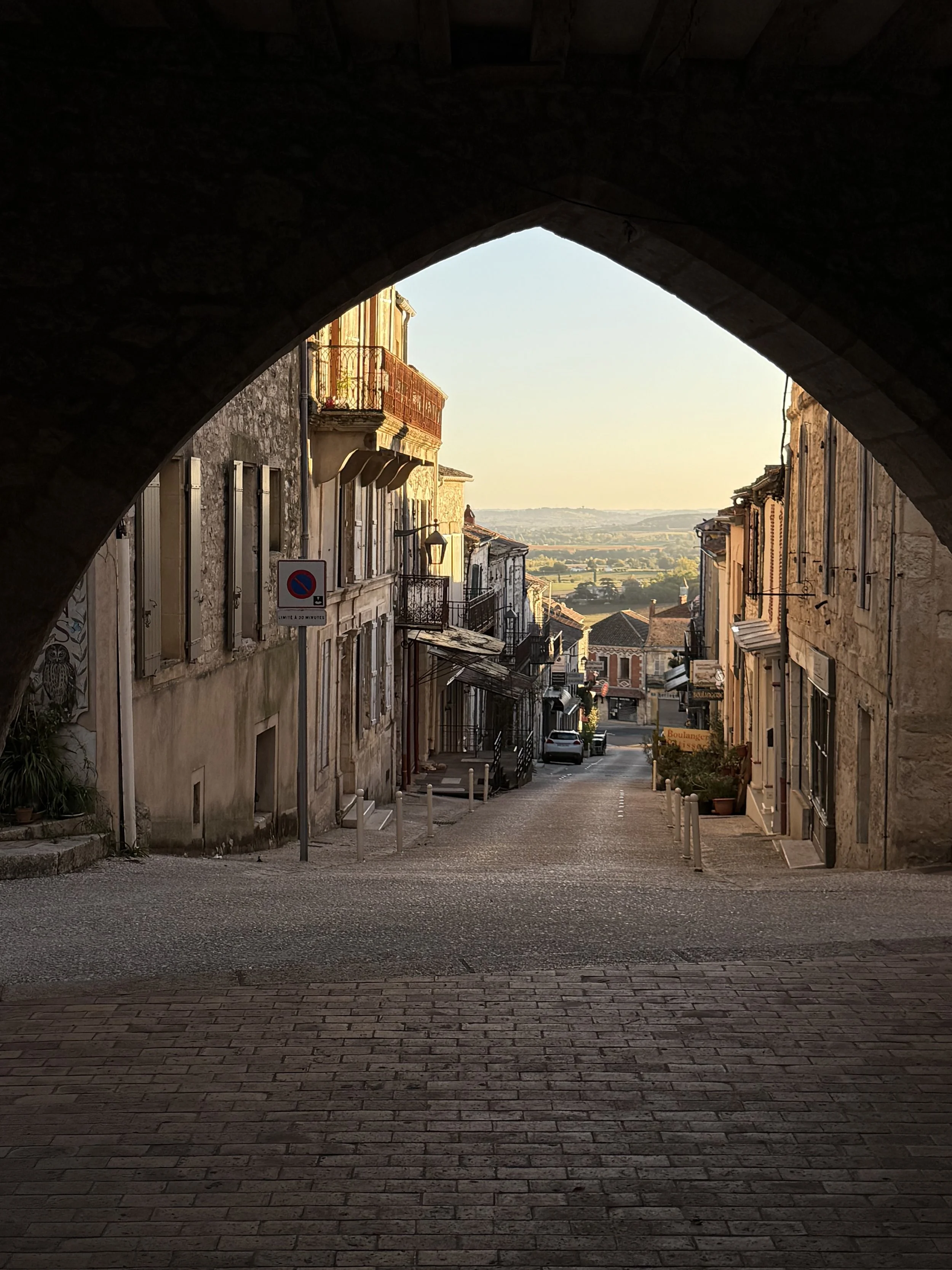 View through a dark archway of a European street with old stone buildings, balconies, and a distant countryside landscape at sunset.