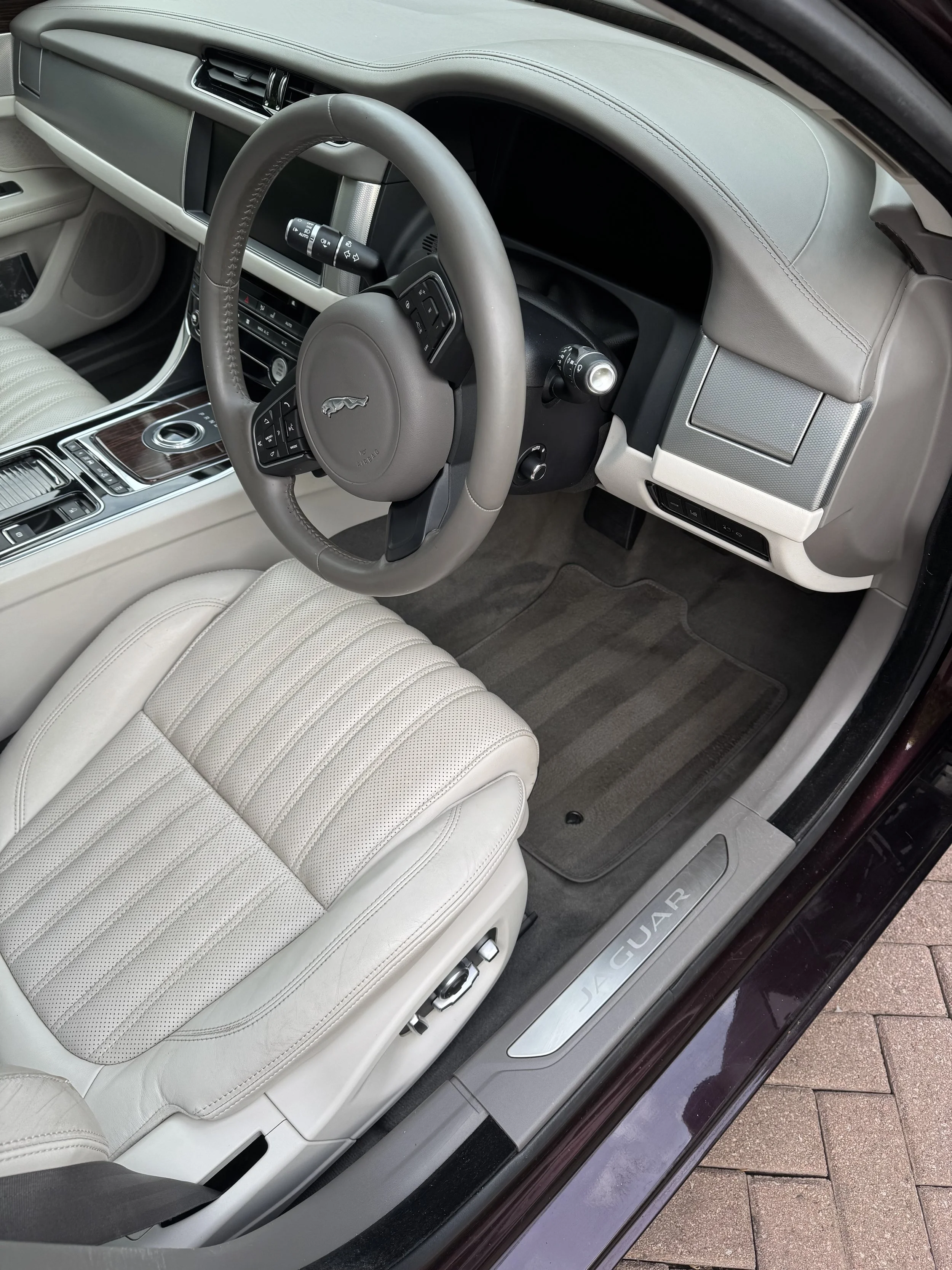 Interior of a luxurious Jaguar car, showing a cream-colored leather seat, grey steering wheel with a Jaguar logo, wood and chrome center console, and a clean beige dashboard with various controls and a digital screen.