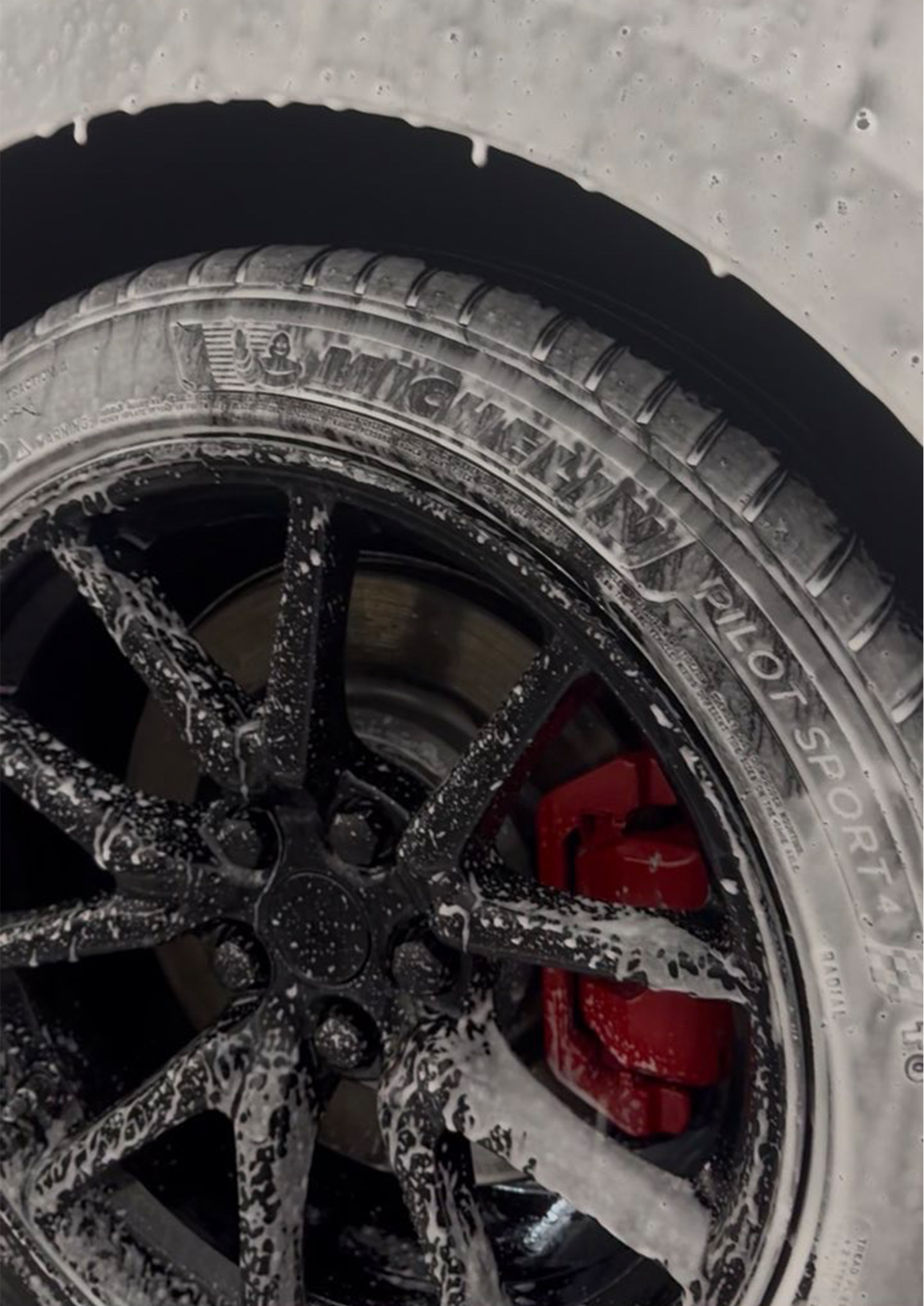 Close-up of a car tire with soap suds and spinning wheel during car wash, with a red object visible in the background.