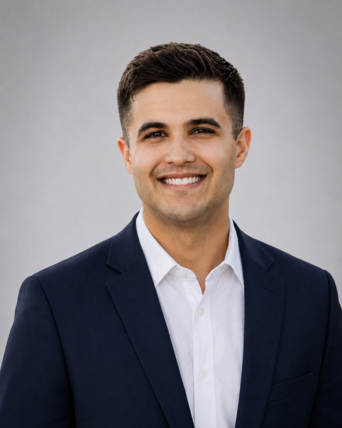 Portrait of a smiling young man wearing a navy blue suit and white shirt against a plain background.