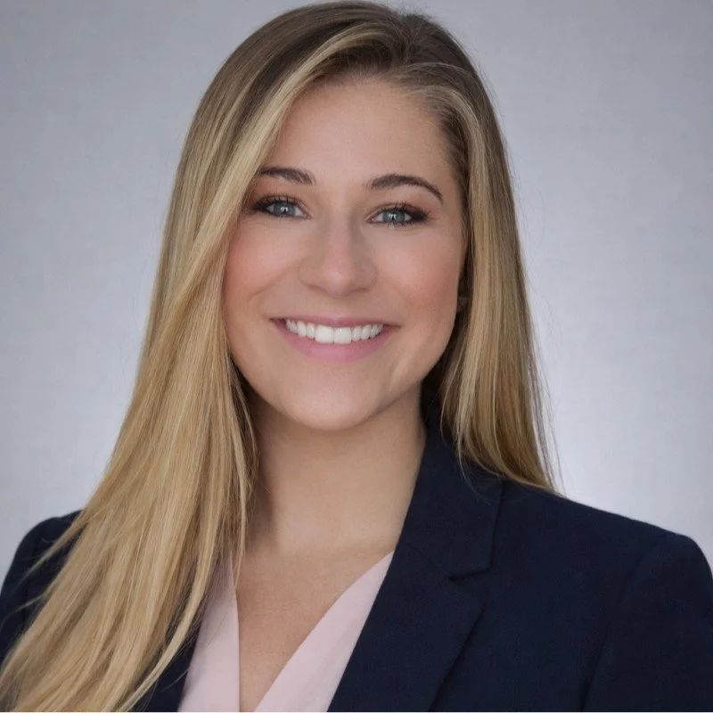 A young woman with long blonde hair and a bright smile, wearing a dark blazer and light-colored top against a neutral background.