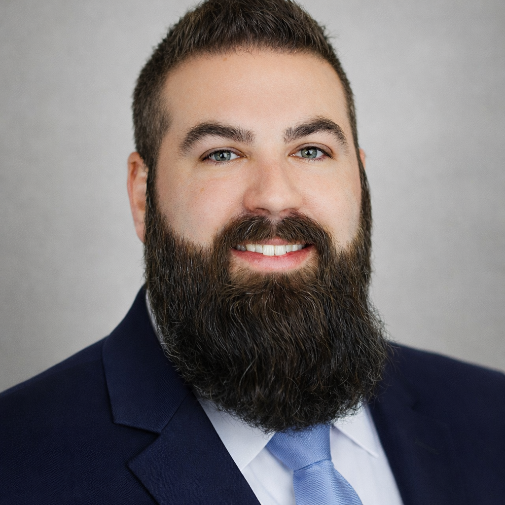 A man with a well-groomed beard and short hair wearing a navy suit, light blue shirt, and matching tie, smiling at the camera.