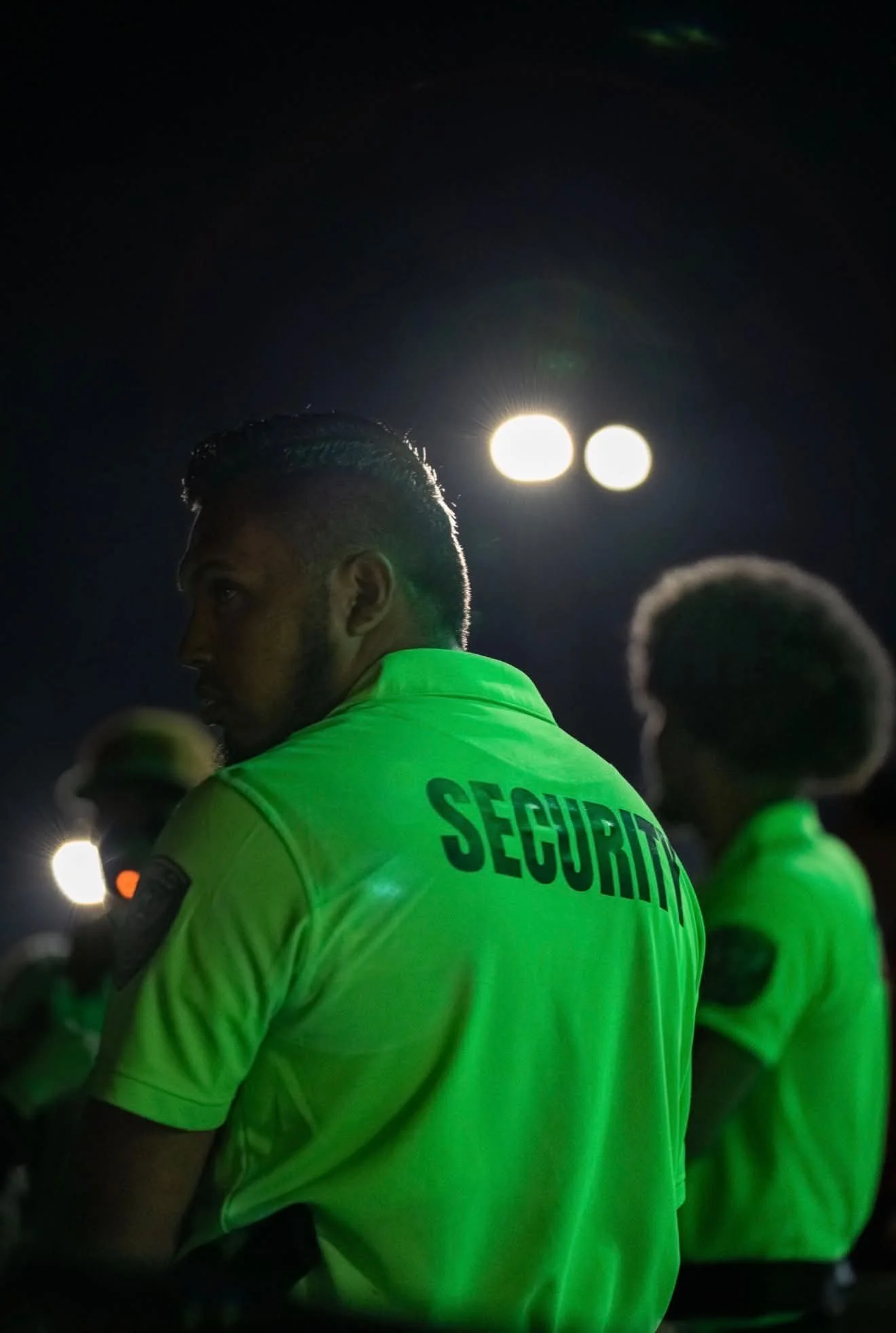 A man wearing a bright green security uniform with the word 'SECURITY' on the back, standing outdoors at night, with a bright light in the background.